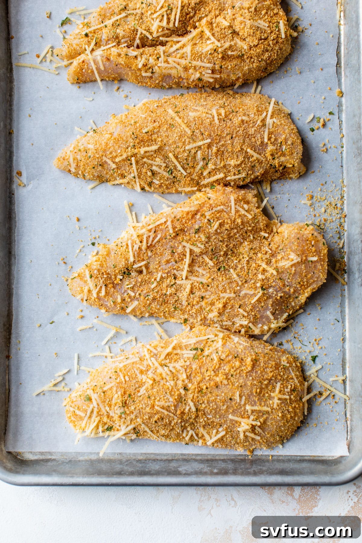 Raw chicken breasts, fully coated in cheese and breadcrumbs, arranged neatly on a wire rack set on a baking sheet, ready for baking.