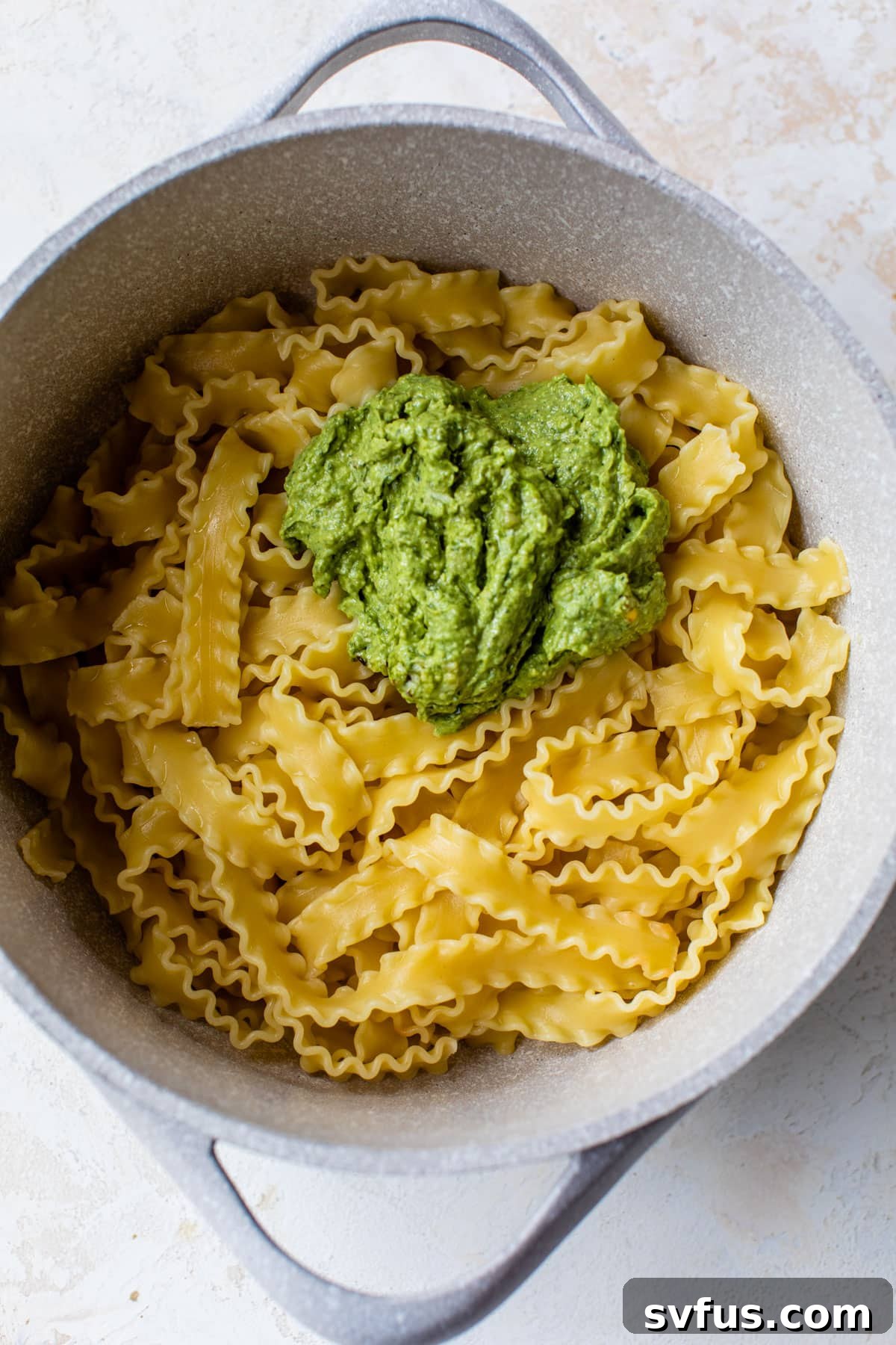 Close-up of freshly cooked pasta noodles inside a large cooking pot, with vibrant green avocado pesto sauce added on top, ready for tossing.