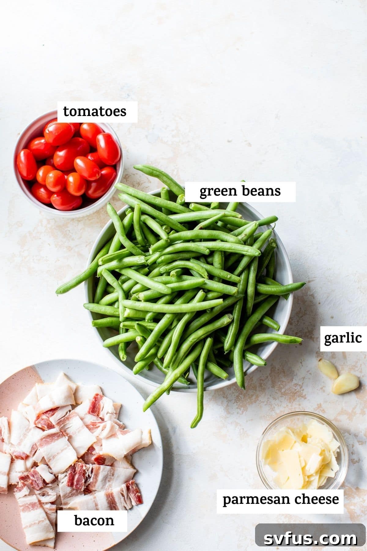 A cutting board with fresh green beans, grape tomatoes, a head of garlic, olive oil, and parmesan cheese, ready to be prepared for the green bean salad.