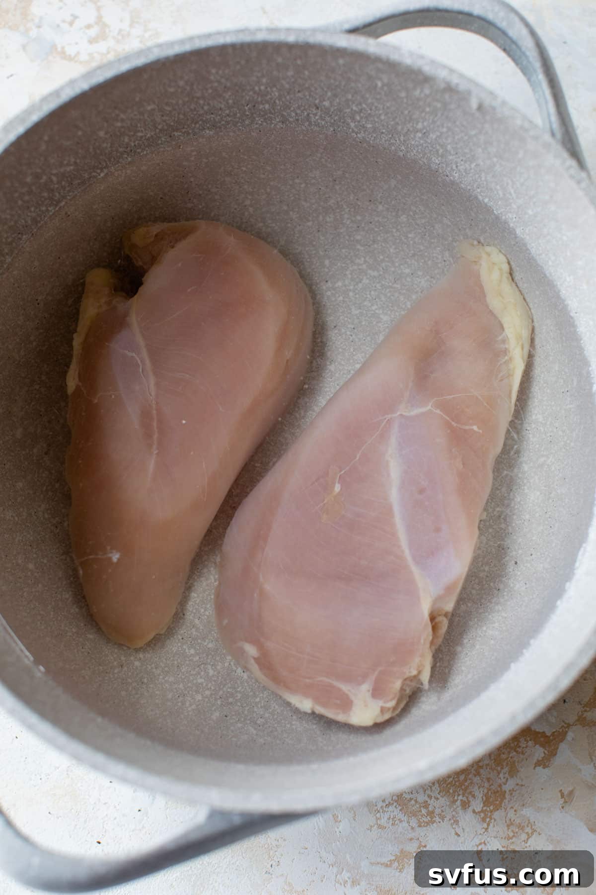 Two chicken breasts submerged in water inside a large pot on the stovetop.