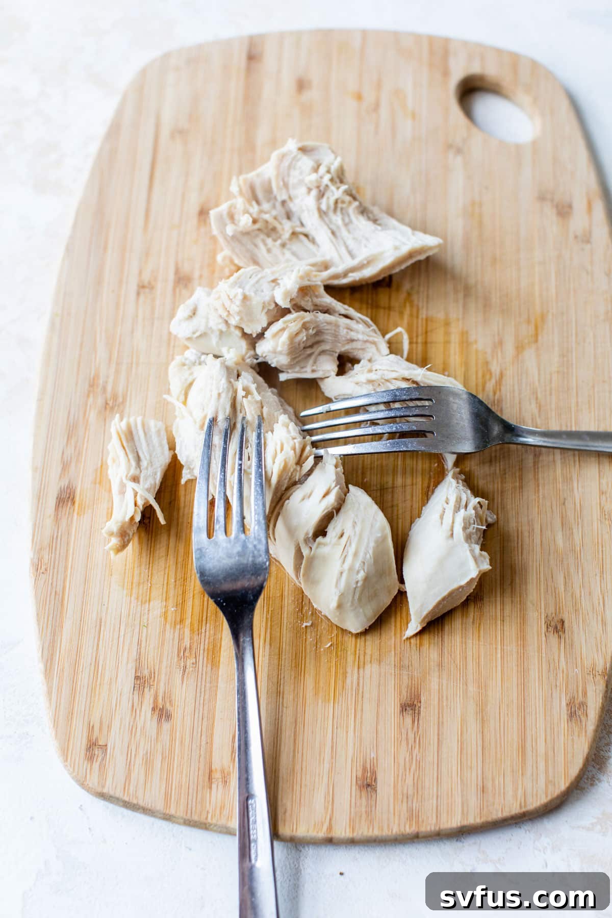 Cooked chicken being shredded with two forks on a wooden cutting board.