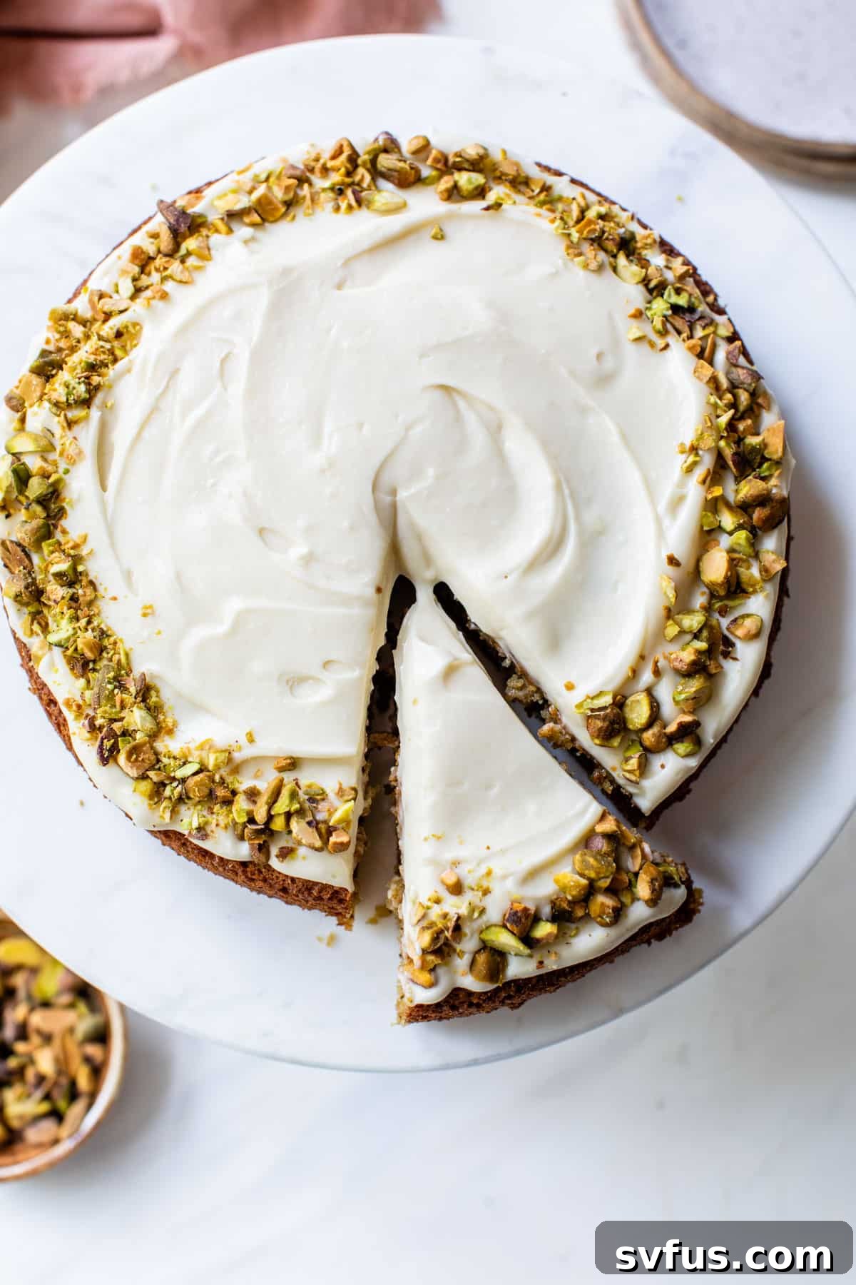 A round, naturally green pistachio cake with a slice removed, revealing its moist interior, topped generously with creamy mascarpone frosting and chopped pistachios.