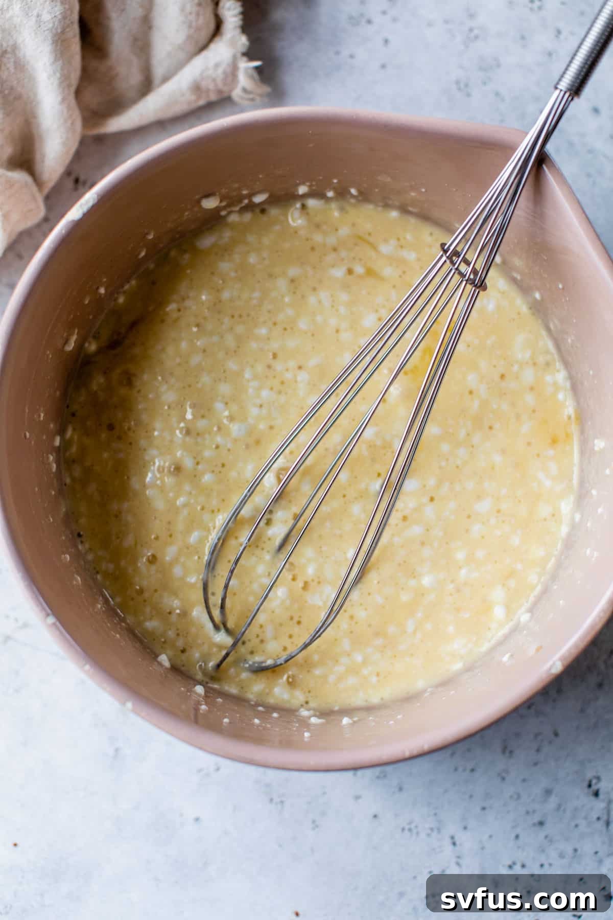 A large bowl filled with wet ingredients for pancakes, including eggs, milk, maple syrup, cottage cheese, and vanilla, being thoroughly whisked together.