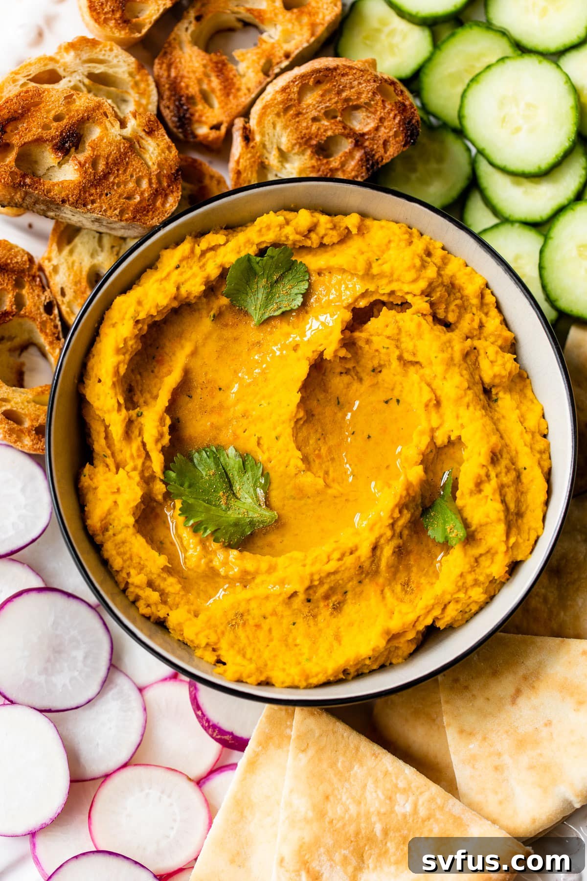 An overhead view of a beautifully garnished bowl of carrot hummus, surrounded by fresh pita bread and colorful vegetables for dipping.