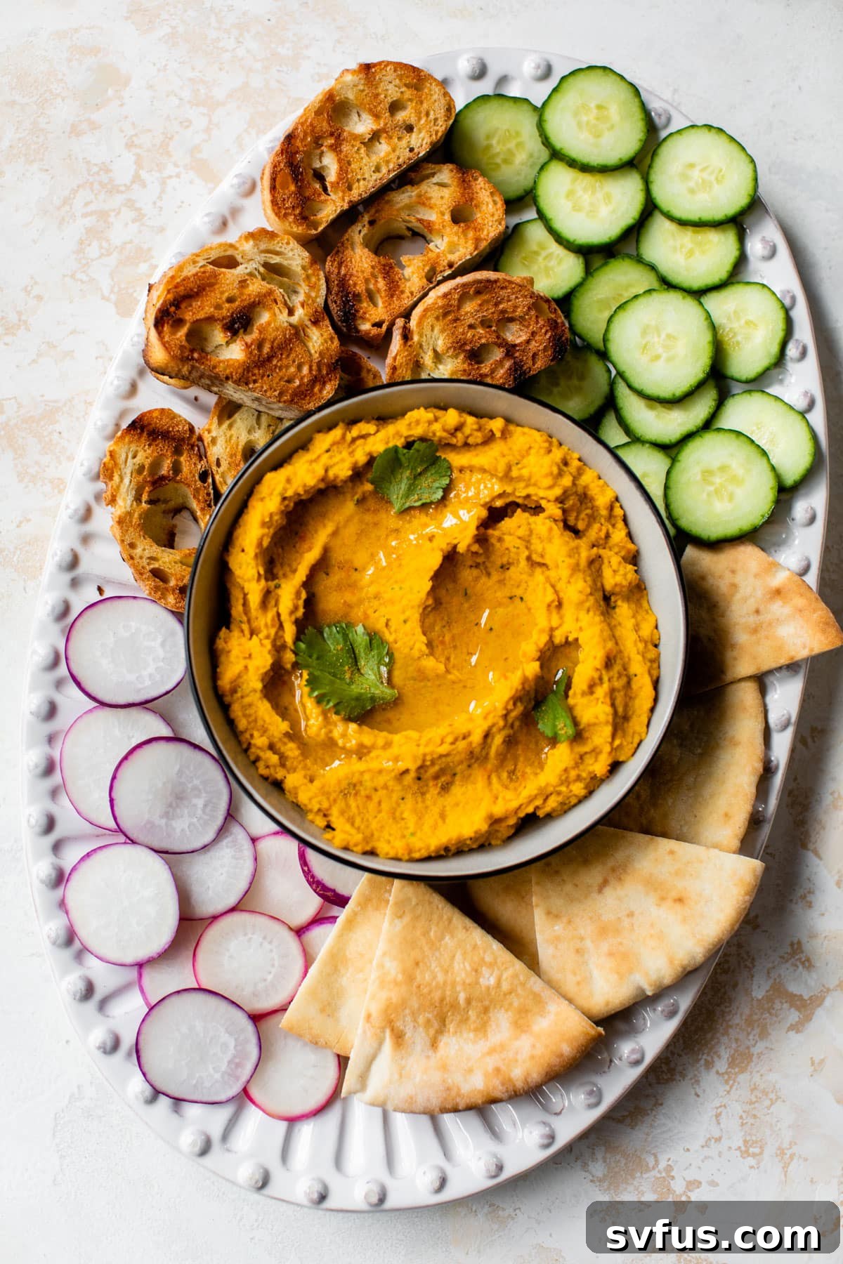 An aerial shot of a beautifully arranged platter featuring a bowl of carrot hummus, surrounded by a variety of colorful fresh vegetables and warm pita bread.