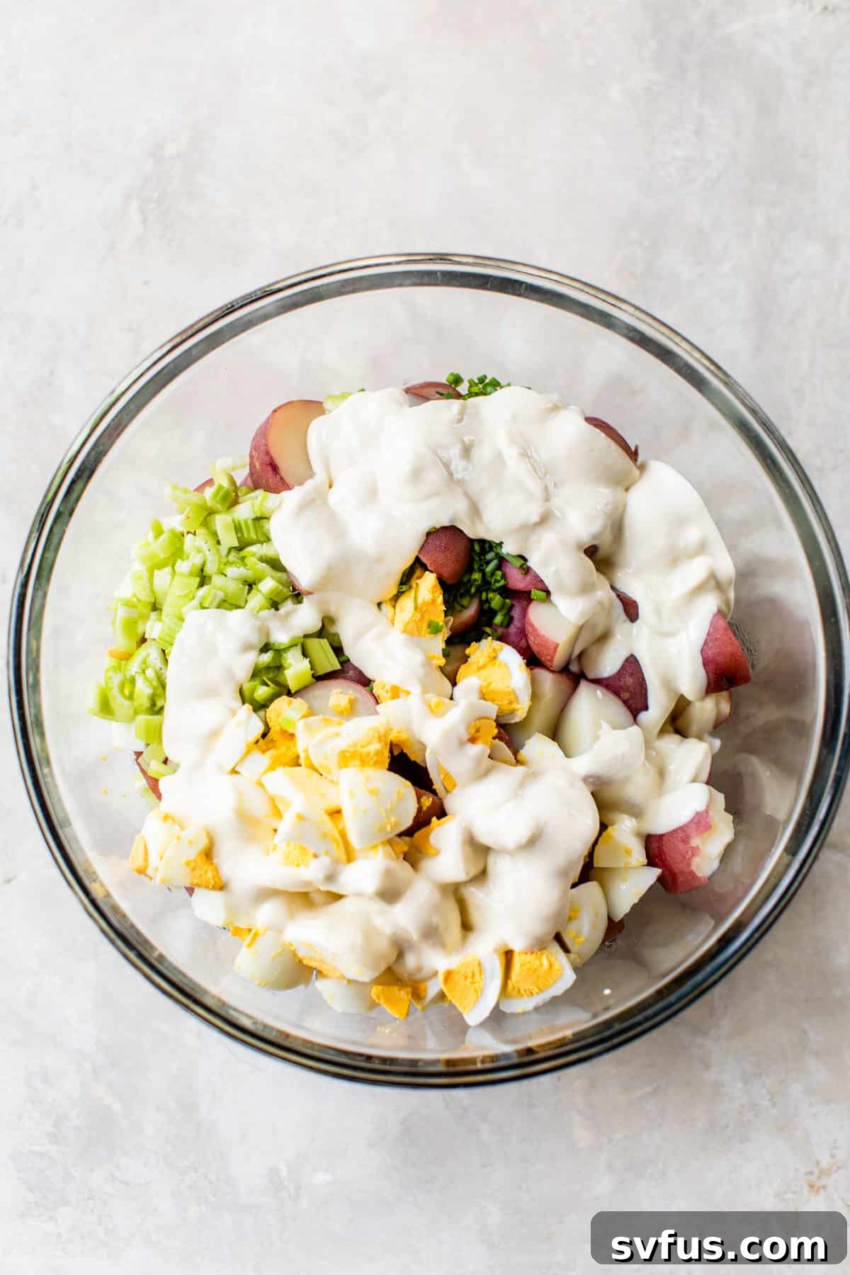 Healthy potato salad ingredients in a large glass bowl with the creamy Greek yogurt dressing being poured over the top, ready for tossing.
