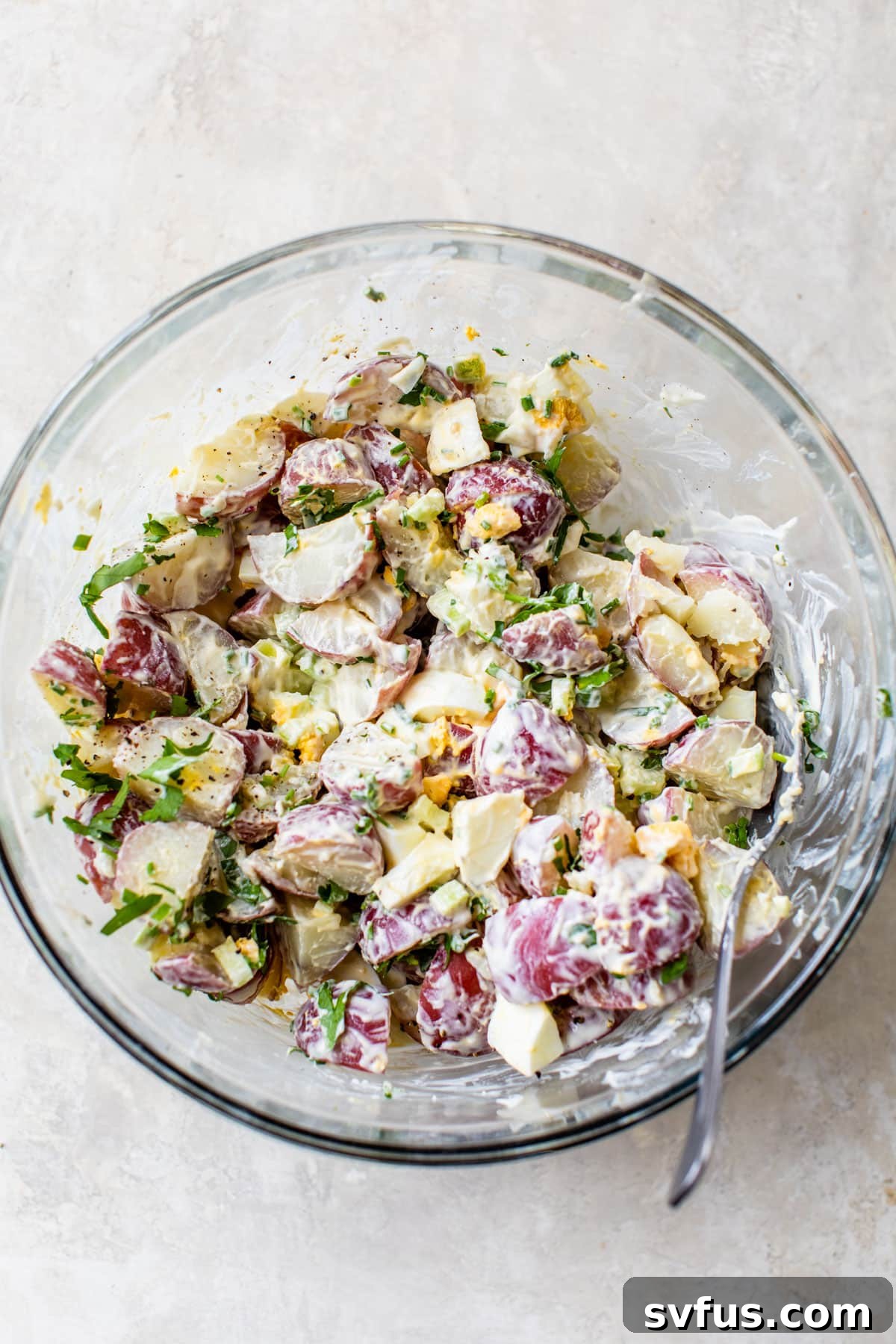 Healthy potato salad ingredients being gently mixed together in a glass bowl to ensure even coating of the dressing.