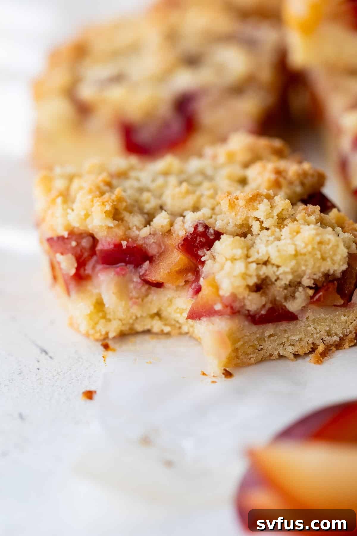 A close-up view of a perfectly baked plum bar, showing its juicy plum filling and golden crumb topping, with a bite taken out.