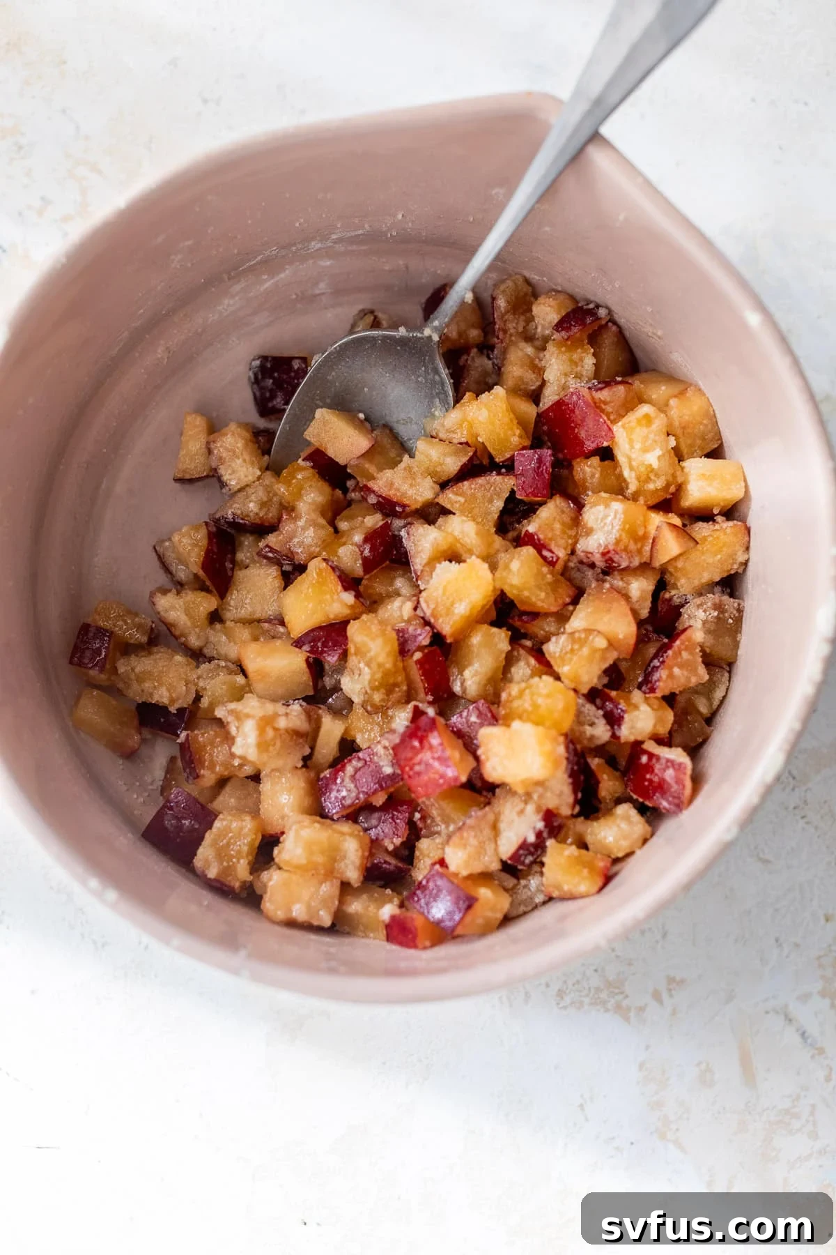 A spoon actively stirring fresh plum chunks with sugar, flour, and vanilla extract in a vibrant pink bowl, preparing the filling for plum bars.