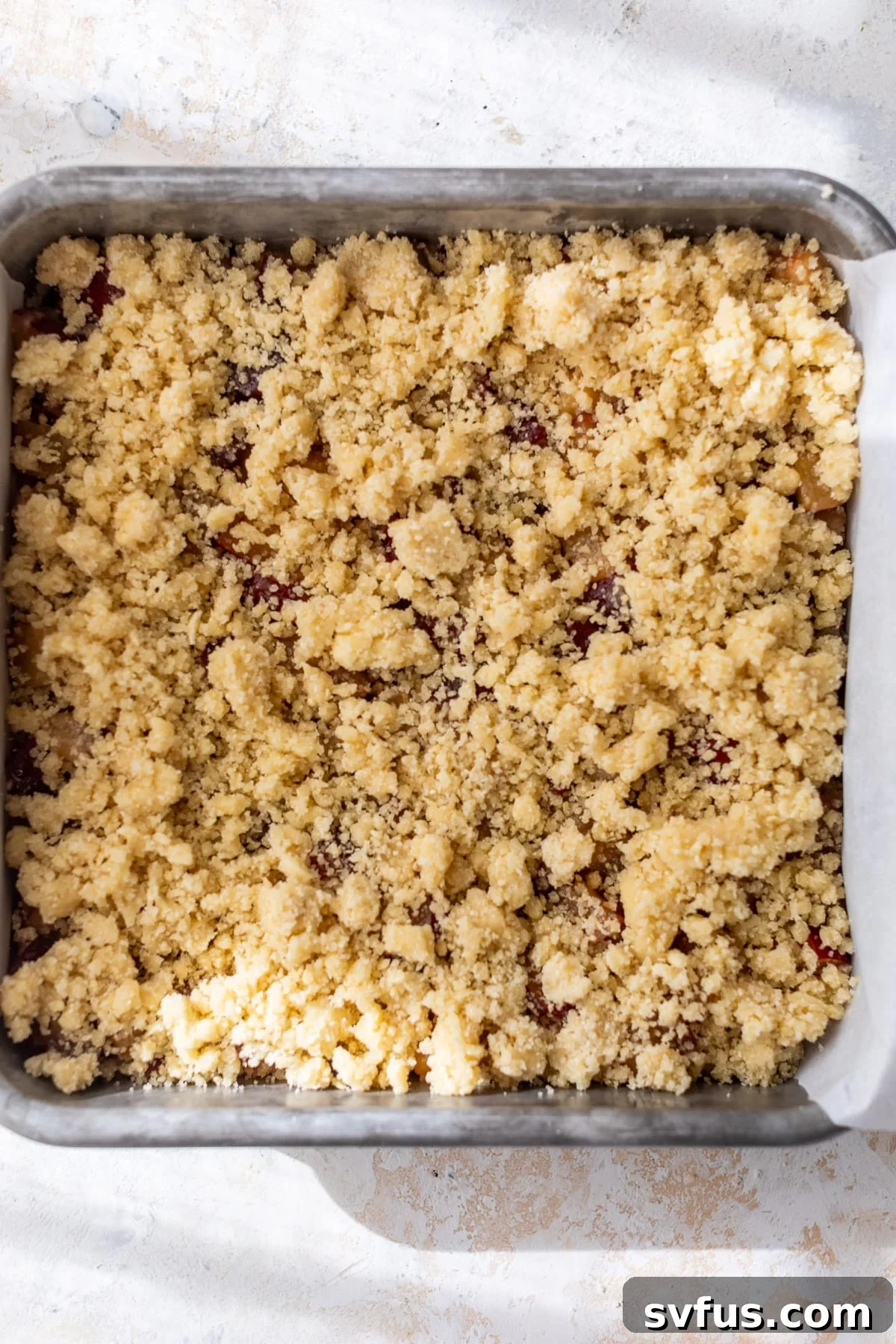 An assembled unbaked plum crumb bar in a baking dish, showing the base crust, plum filling, and crumb topping before baking.