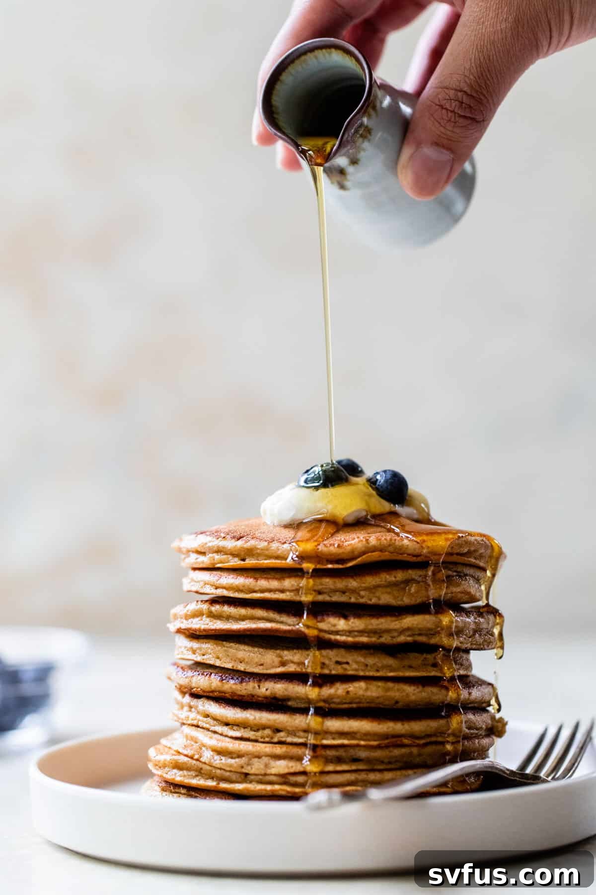 maple syrup pouring over a stack of pancakes with yogurt and blueberries on top