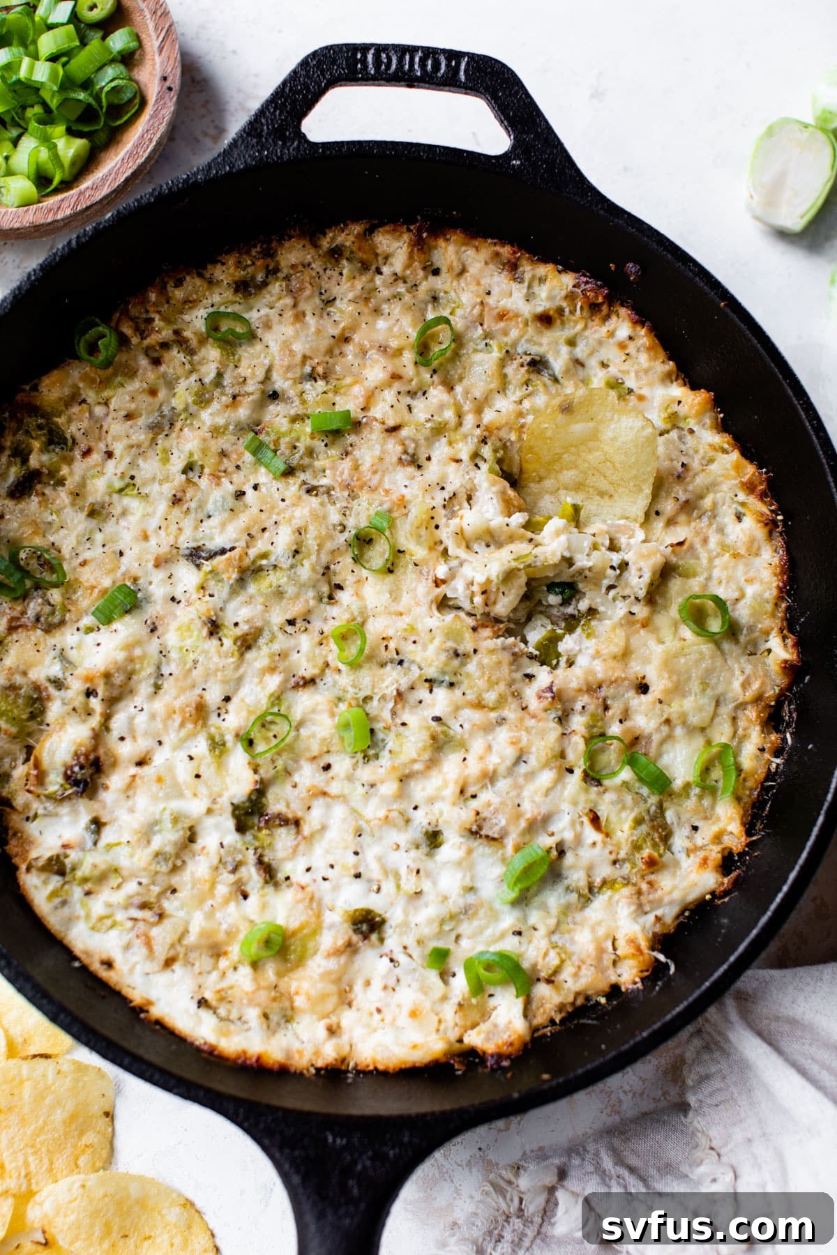 Close-up of baked Brussels Sprouts Dip in a cast iron skillet, with a single potato chip ready for dipping