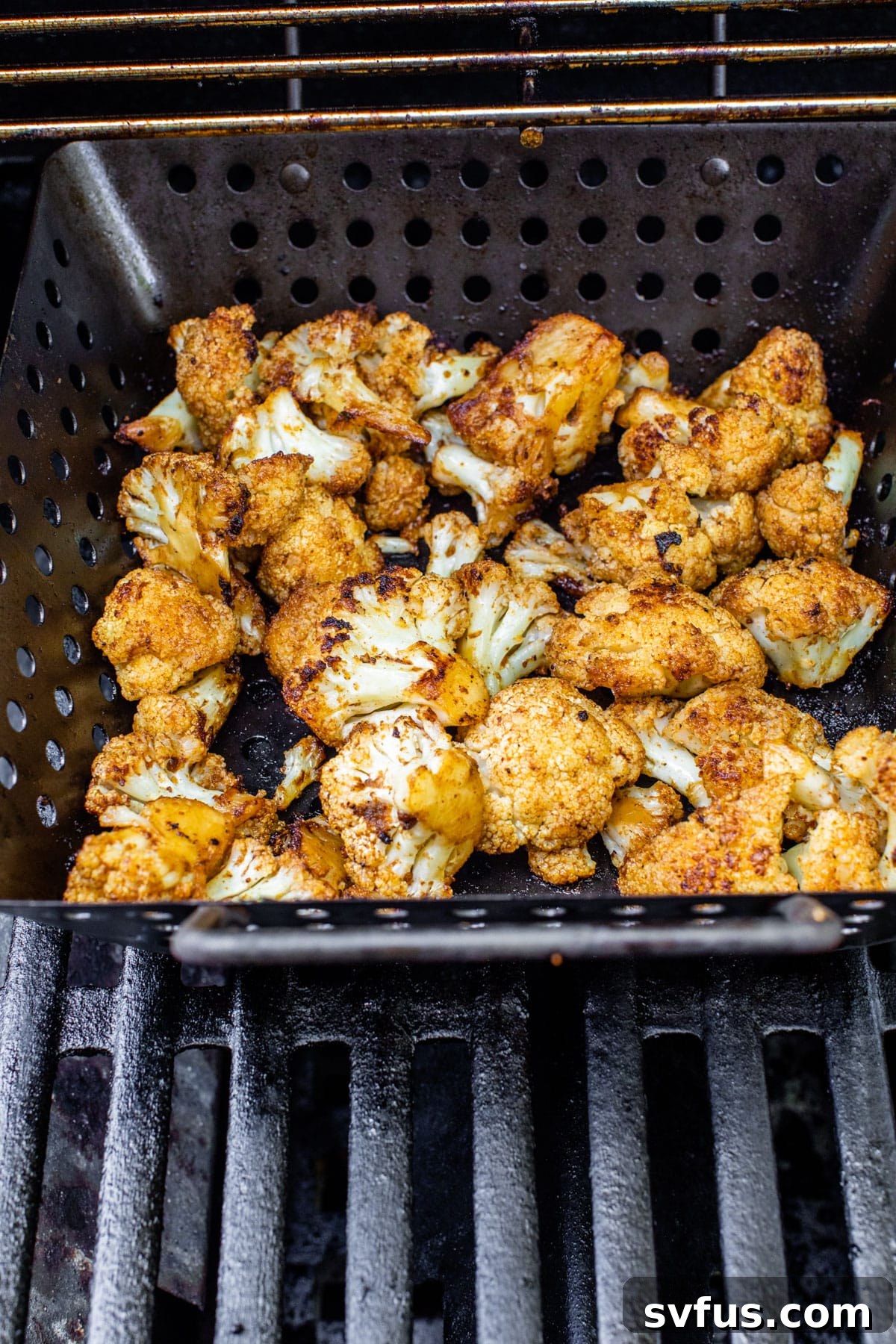 Spicy grilled cauliflower florets cooking in a metal grill basket on an outdoor grill
