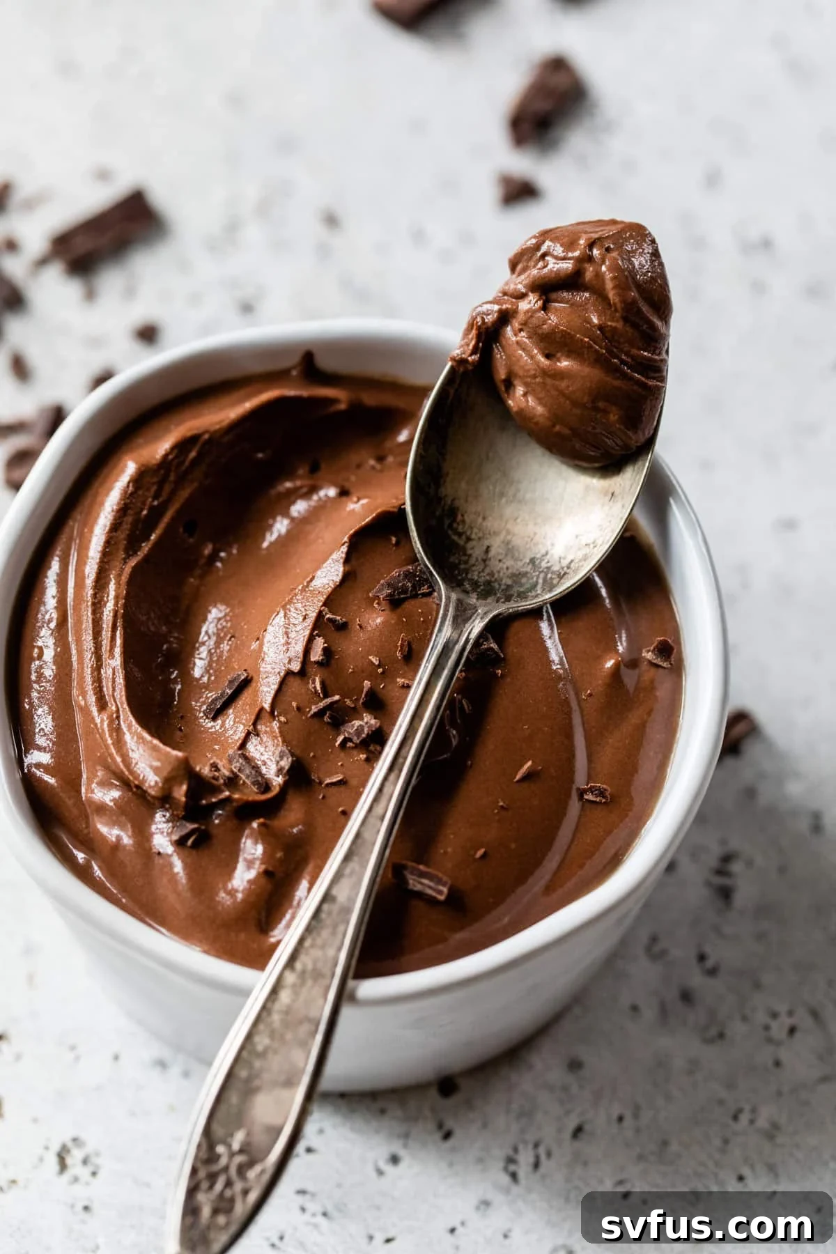 A glistening spoonful of smooth vegan chocolate mousse hovering above a small, elegant bowl filled with the same creamy dessert, hinting at its luxurious texture.