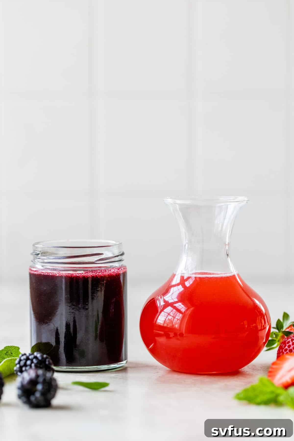 Two glass jars filled with different colored simple syrups, one dark (blackberry) and one lighter (grapefruit), symbolizing the two distinct shrub flavors.