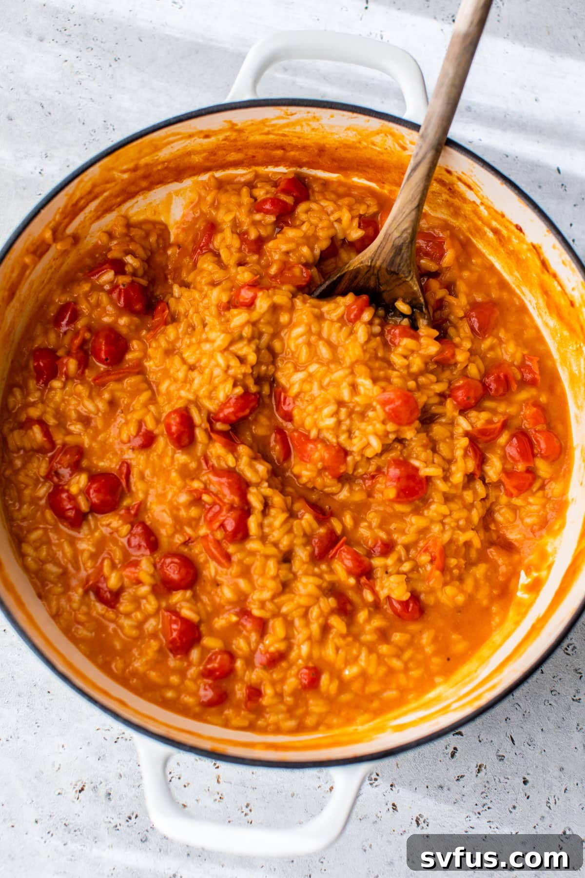 A large, deep skillet brimming with creamy Arborio rice, glistening grape tomatoes, and a wooden spoon resting in the dish.