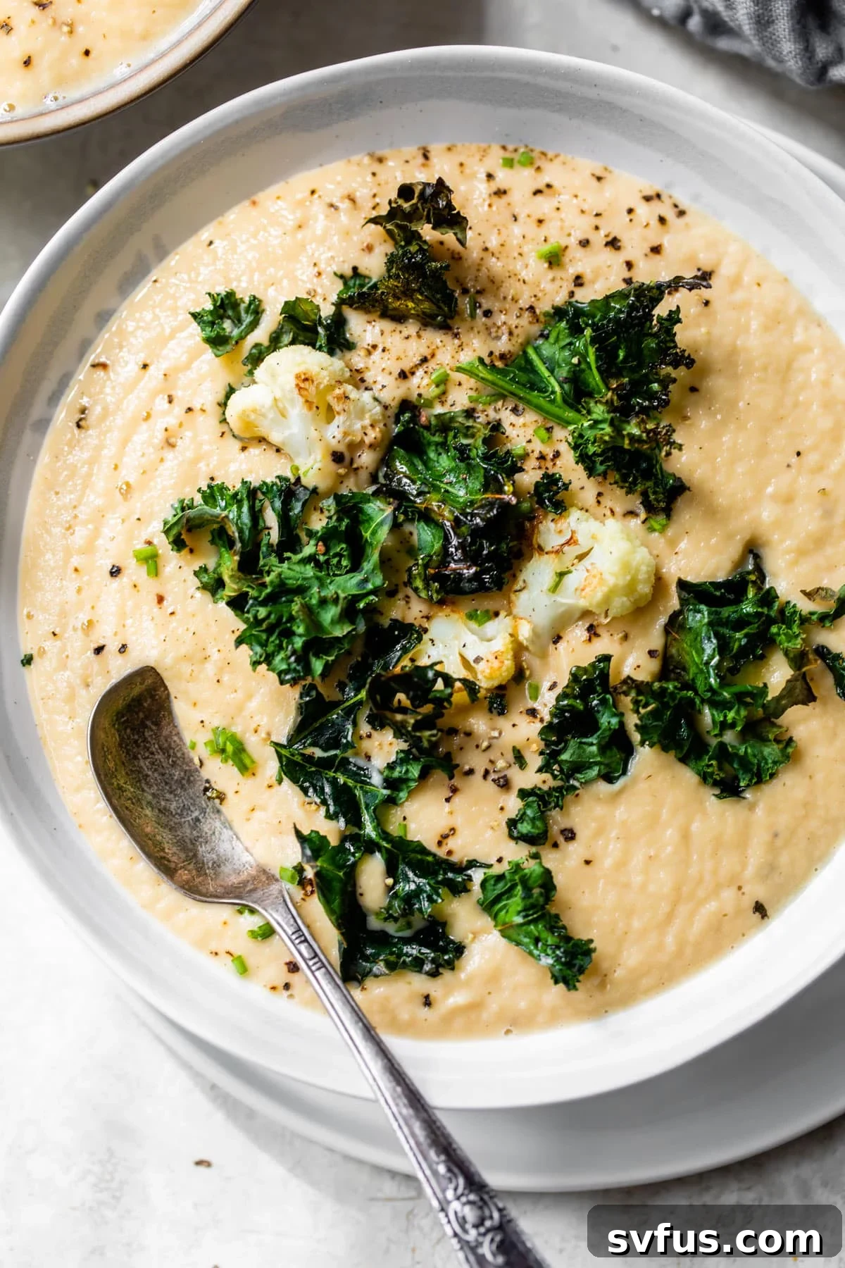 A beautifully presented bowl of creamy, golden-yellow vegan cauliflower soup, garnished with delicate roasted cauliflower florets and vibrant green kale chips.