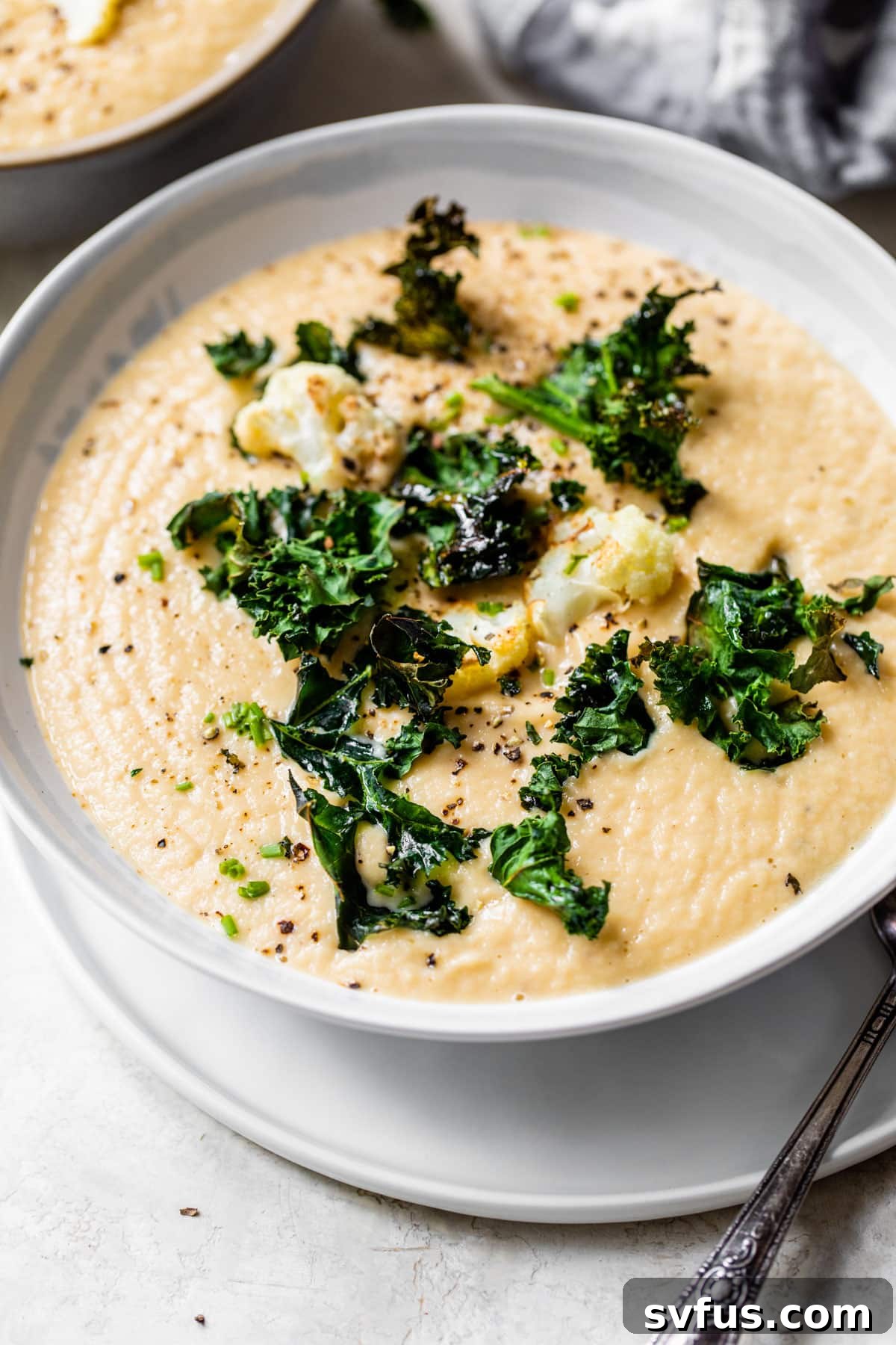 A beautifully presented bowl of blended vegan cauliflower soup, topped generously with roasted cauliflower florets and vibrant green kale chips, ready to be enjoyed.
