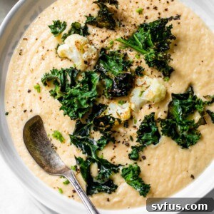 A bowl of creamy, golden-yellow vegan cauliflower soup, garnished with roasted cauliflower florets and vibrant green kale chips.