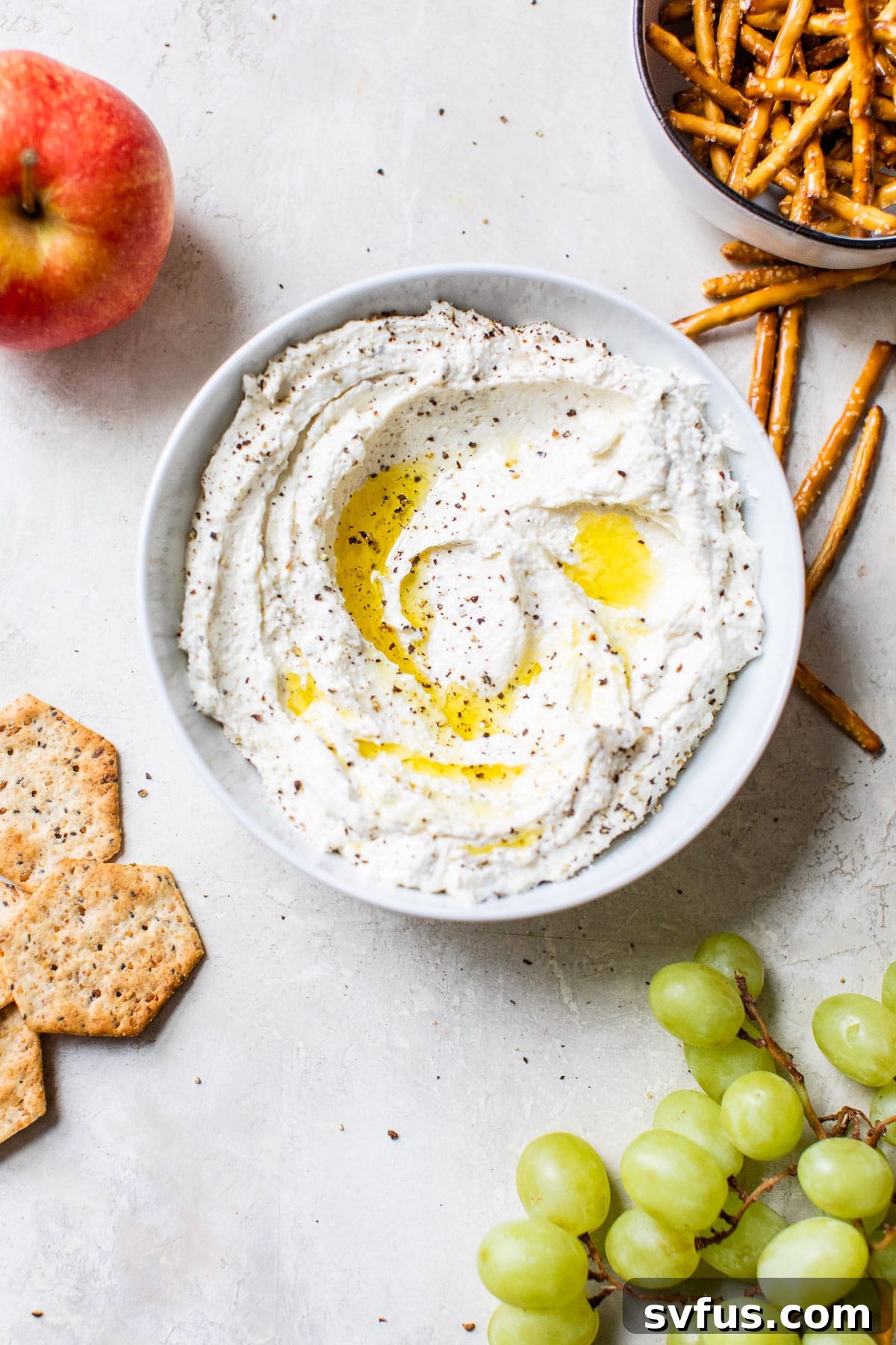 A bowl of creamy black pepper whipped feta surrounded by an assortment of crackers, pretzels, and fresh grapes, perfect for an appetizer spread.