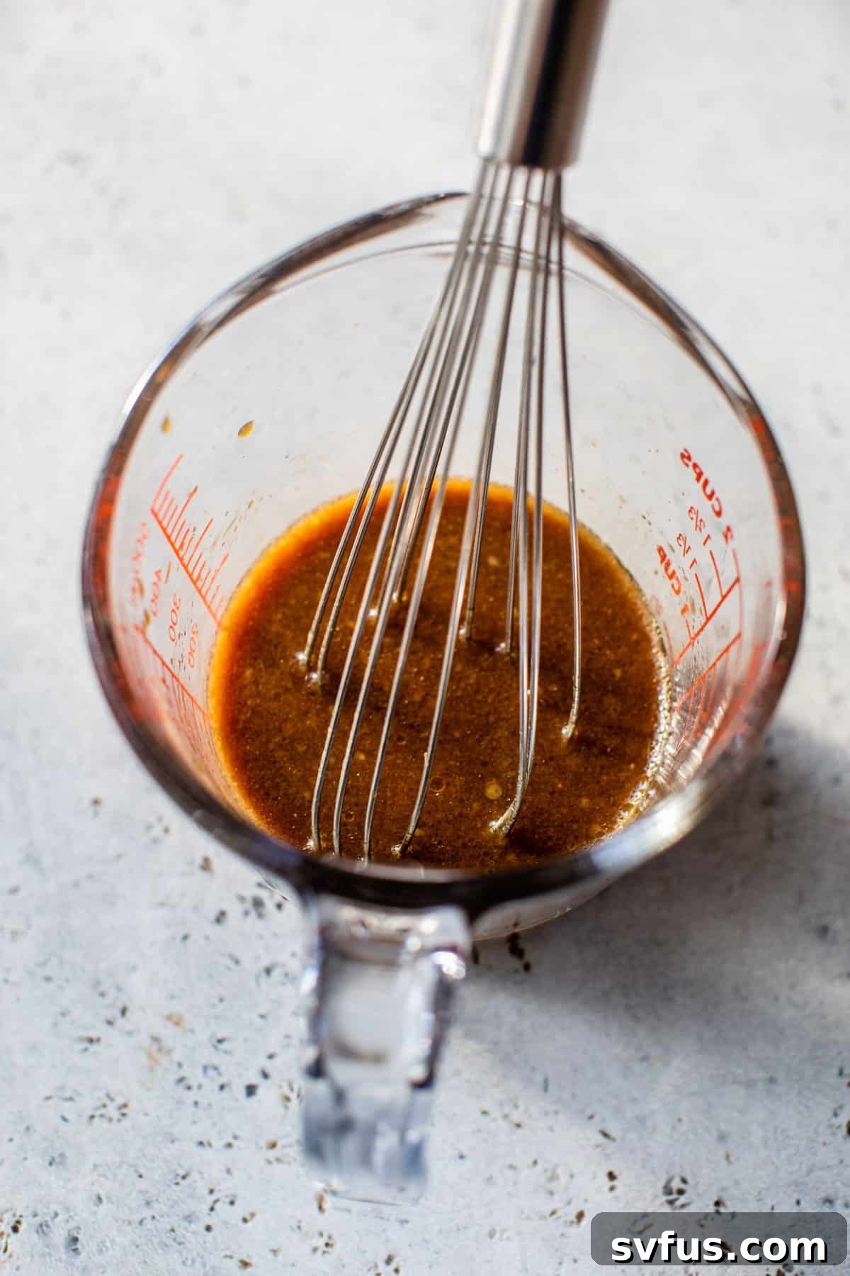 A clear glass measuring cup holding a rich, amber-colored Asian stir-fry sauce, with a small whisk resting inside.