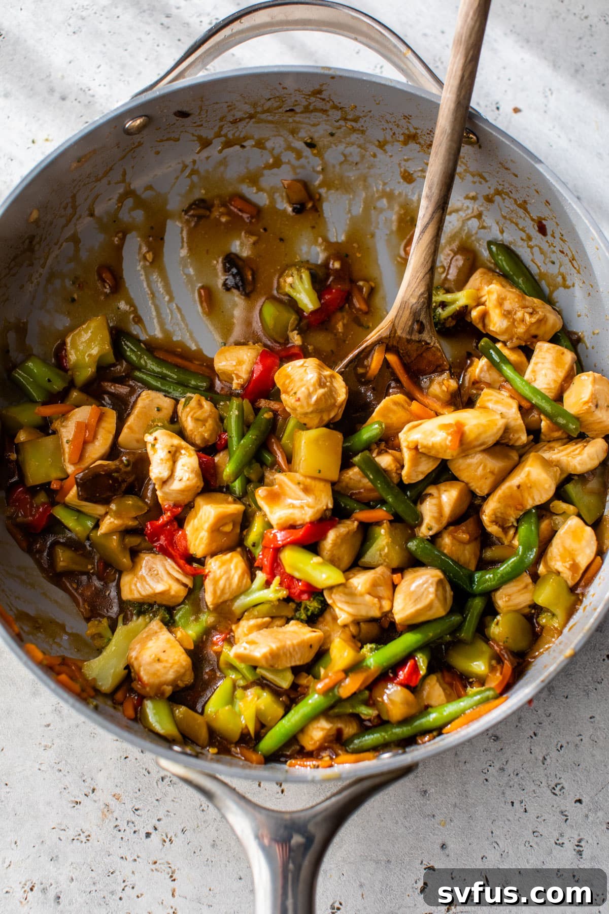 A large skillet filled with chicken and vegetables coated in a rich, thickened stir-fry sauce, with a wooden spoon mixing the ingredients.