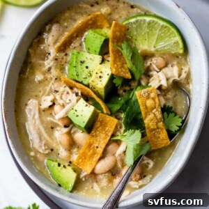 A bowl of soup on a plate with a spoon made with white beans and shredded chicken and topped with diced avocado, tortilla strips and cilantro