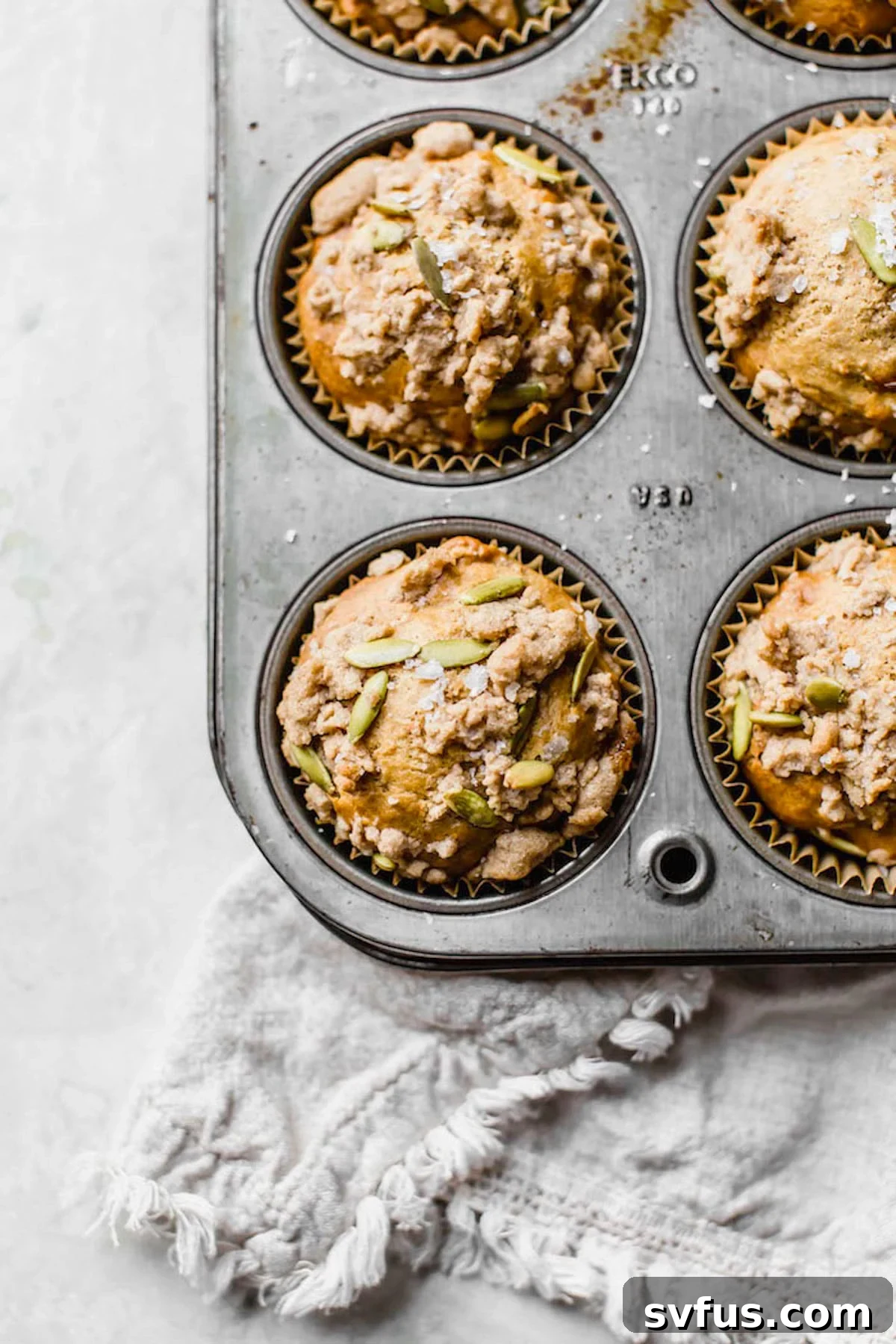 A batch of golden-brown healthy sweet potato muffins fresh out of the oven, nestled in a muffin tin, topped with a crunchy cinnamon pepita crumb.