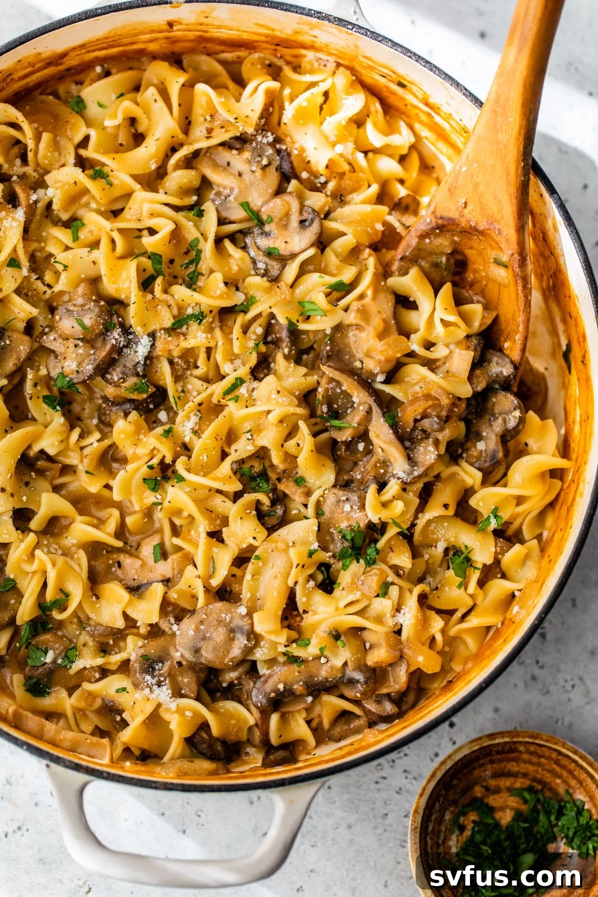 mushroom stroganoff inside a dutch oven with a wooden spoon and fresh parsley and cheese on top