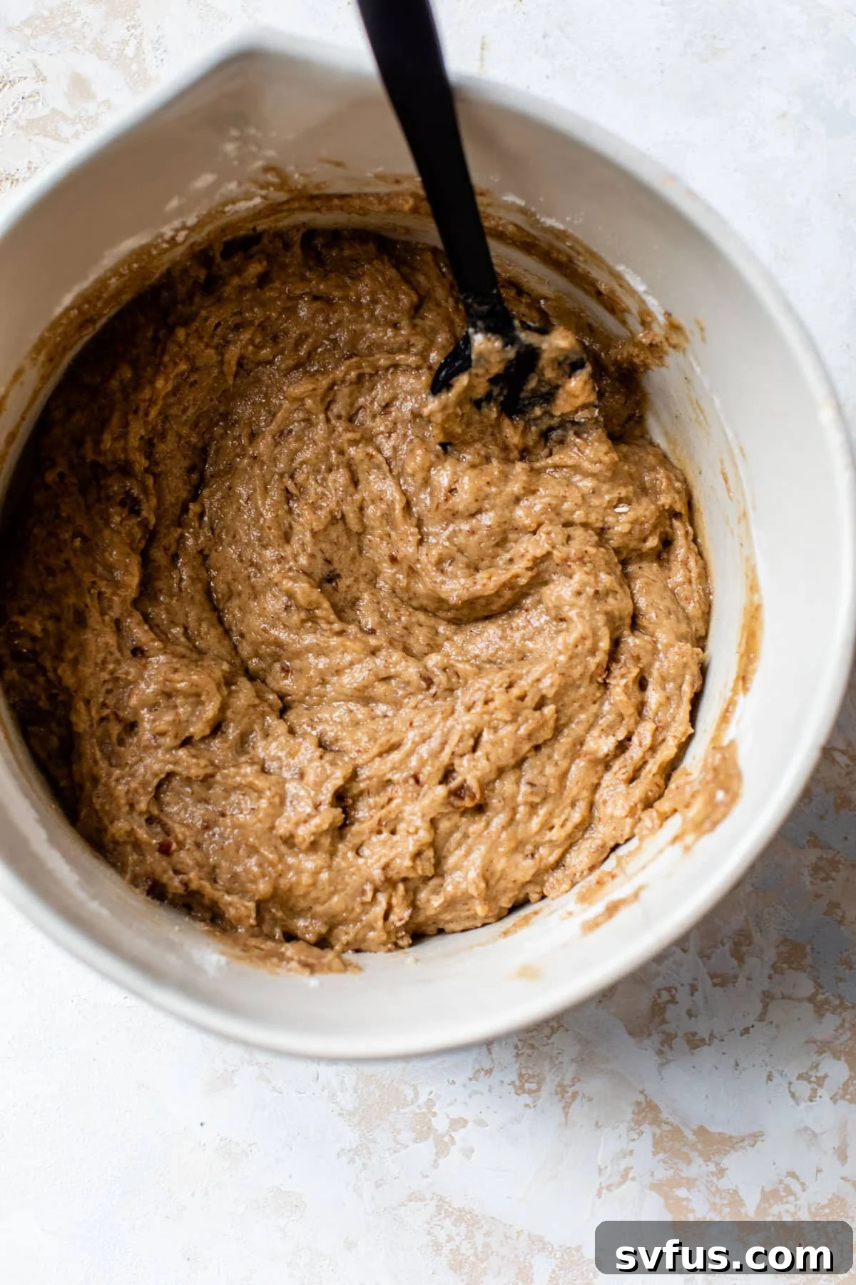 Vegan sticky toffee pudding batter being gently folded with a rubber spatula in a large mixing bowl until just combined, ready for baking.