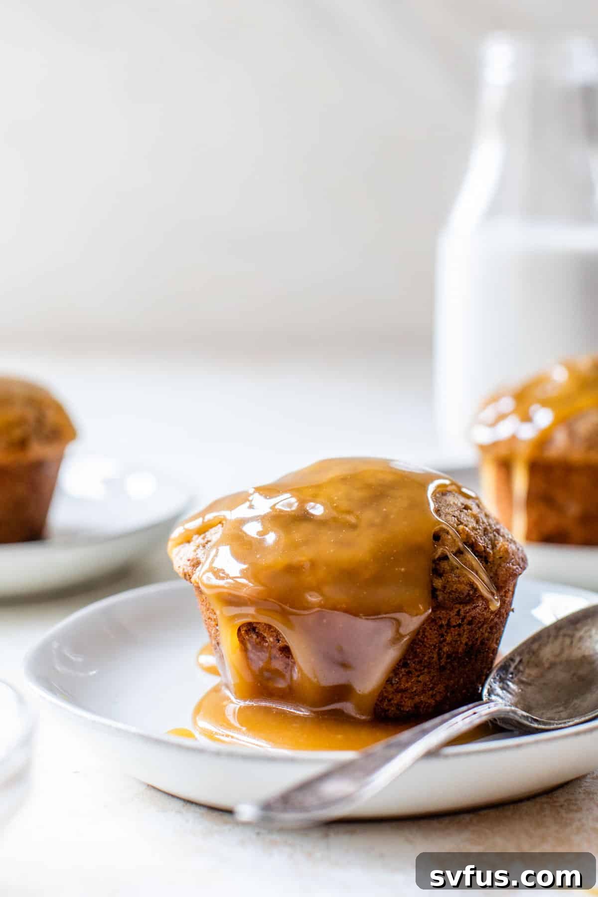 A perfectly assembled vegan sticky toffee pudding on a small white plate, glistening with rich caramel sauce and a small spoon ready for the first bite.