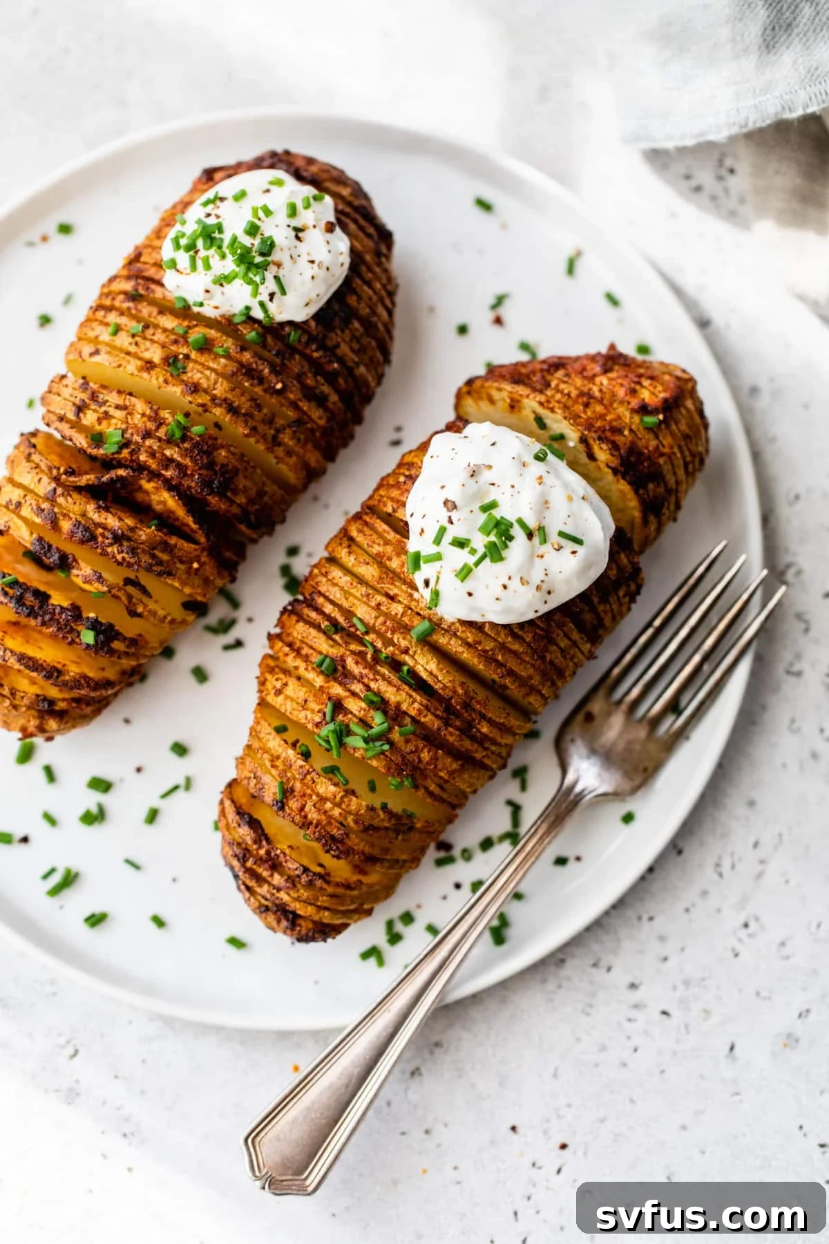 Two air-fried hasselback potatoes, beautifully fanned out and topped with a dollop of sour cream and fresh chives, served on a plate with a fork.