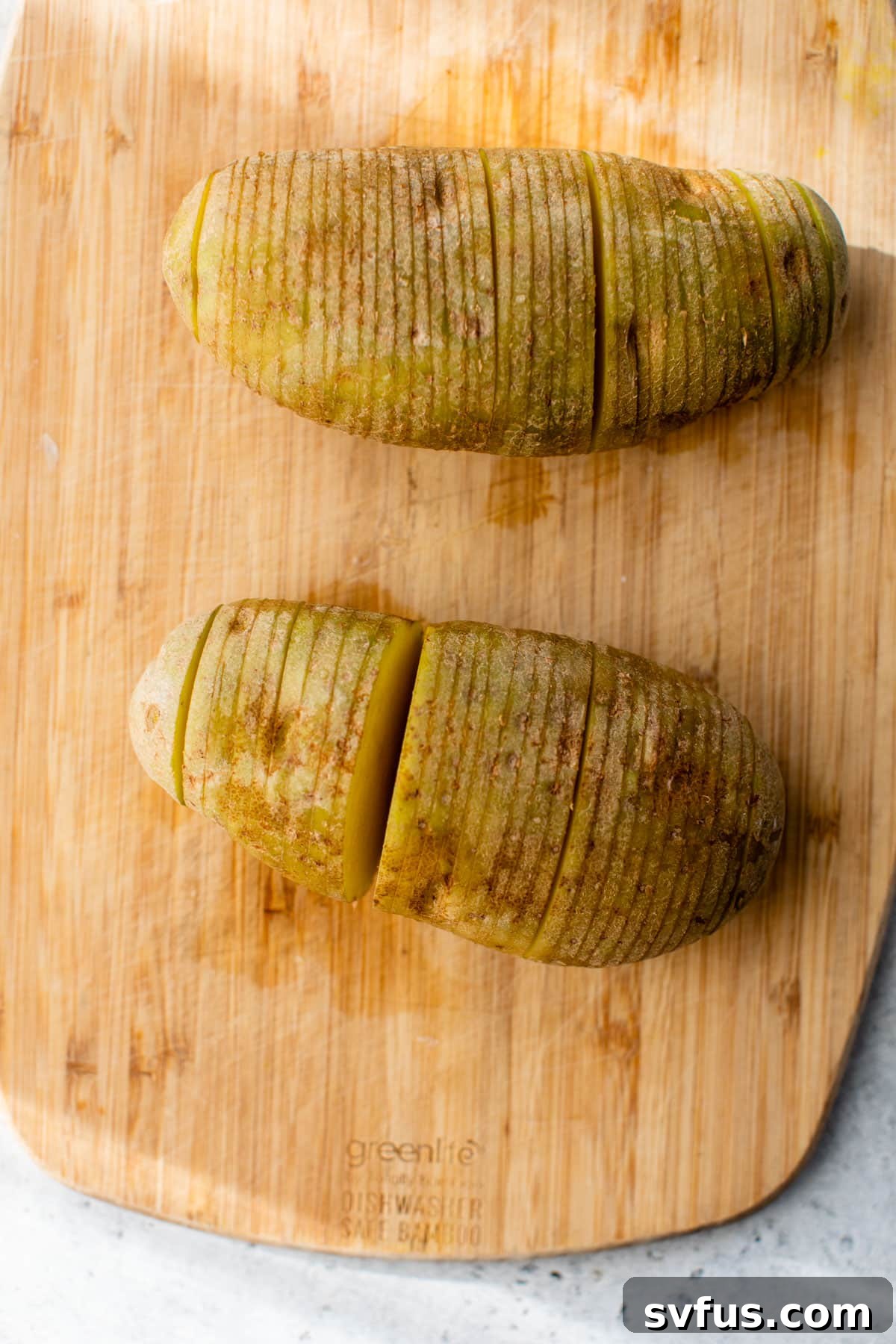 Two Russet potatoes with precise, accordion-style slices on a cutting board, ready for seasoning.