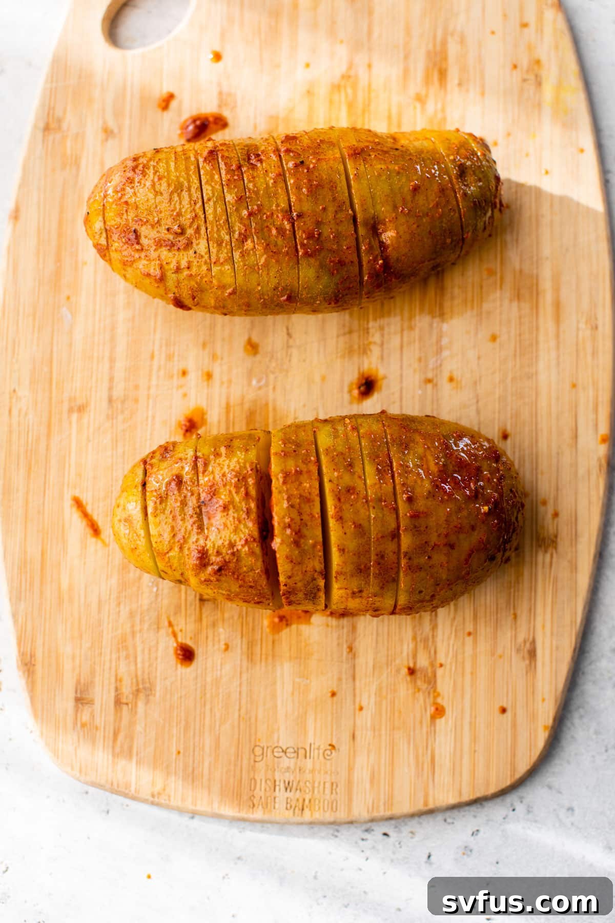 Two sliced potatoes on a cutting board, generously drizzled with melted butter and spice mixture, showing how the seasoning penetrates the cuts.
