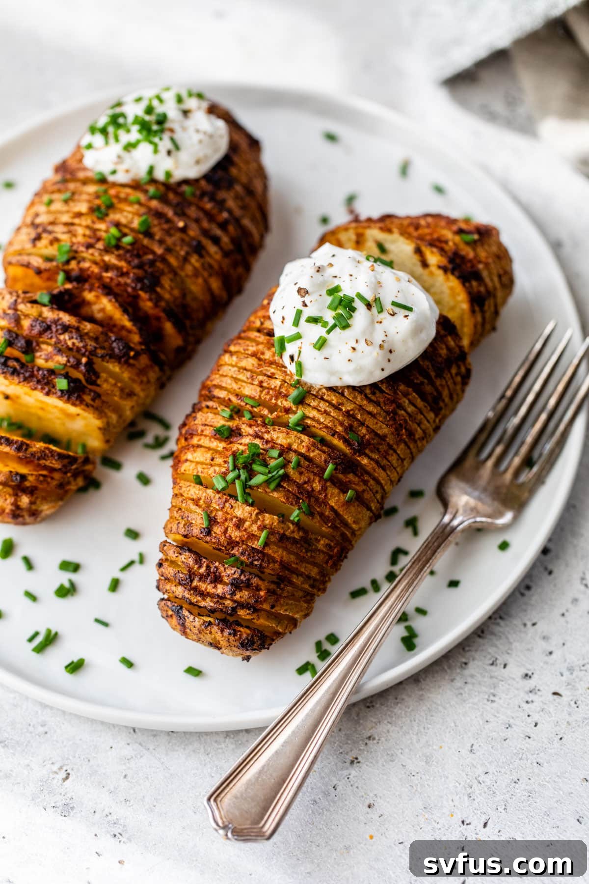Two beautifully presented hasselback potatoes topped with sour cream and chives, served on a plate with a fork, ready to be enjoyed.