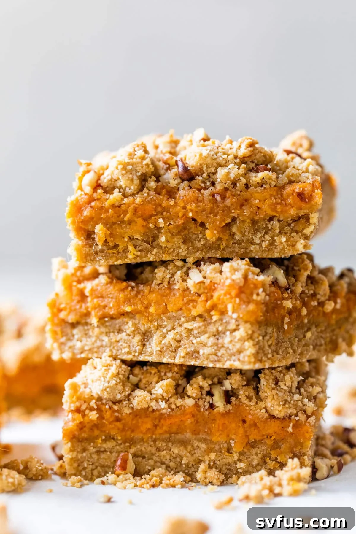 A stack of three golden-brown sweet potato bars with a visible crumb topping, set against a warm, inviting backdrop, emphasizing their deliciousness and layered texture.
