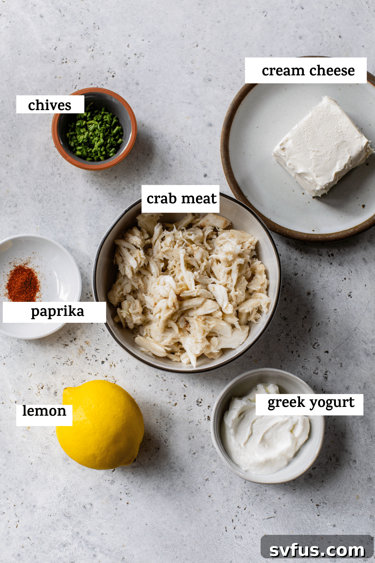 A beautifully arranged selection of fresh ingredients for cold crab dip on a rustic wooden table, including vibrant chives, a block of cream cheese, a small bowl of paprika, tender lump crab meat, a bright lemon, and a container of creamy Greek yogurt.