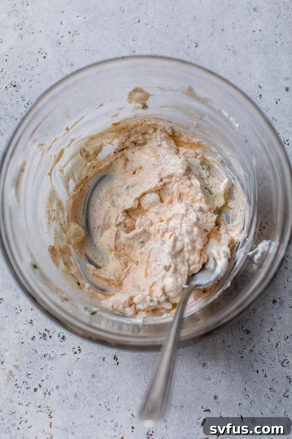 Softened cream cheese, tangy Greek yogurt, and a blend of vibrant paprika and salt are thoroughly combined and whisked together in a clear glass mixing bowl using a spoon, forming a smooth, light orange base for the dip.