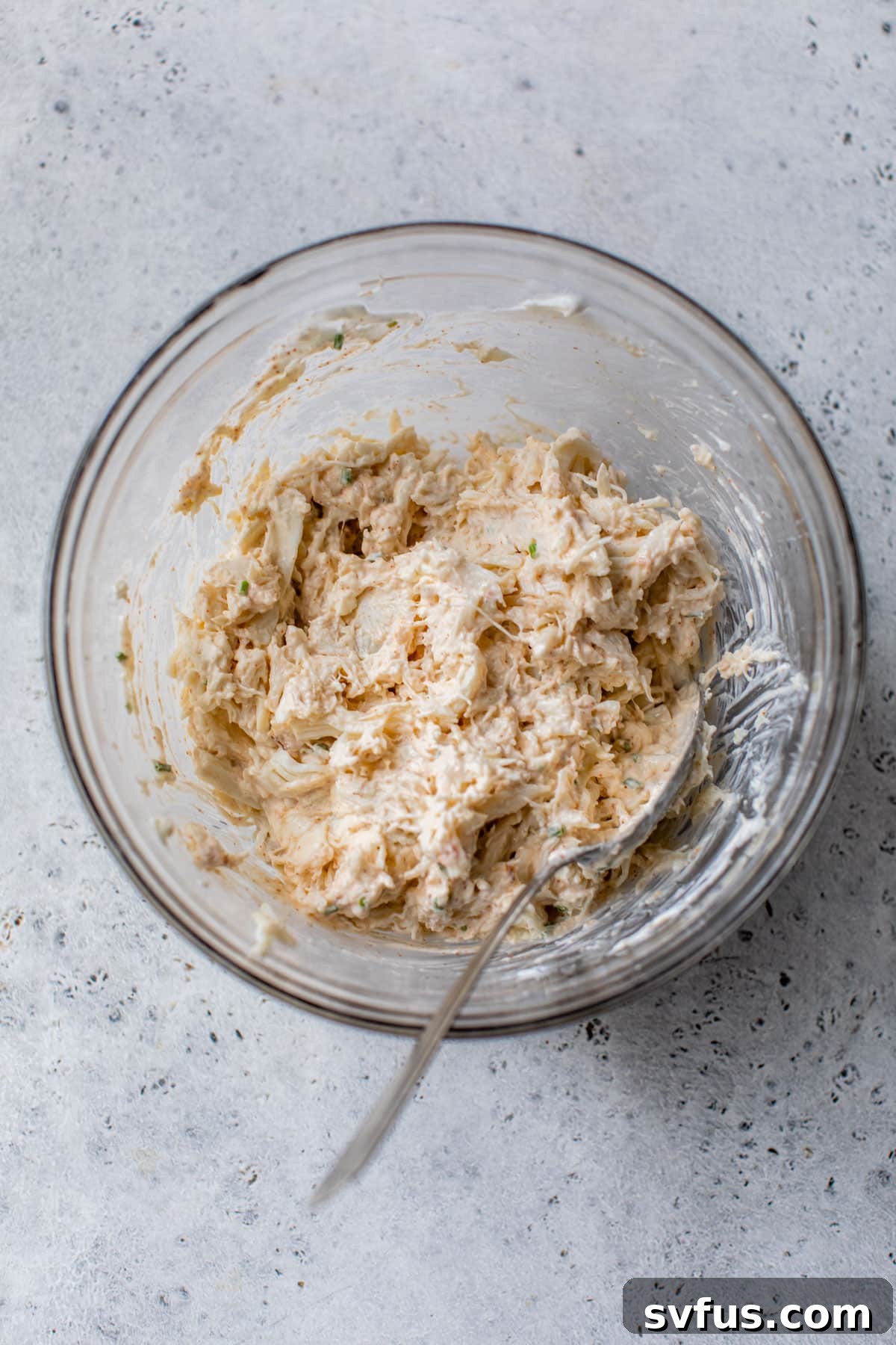 Delicate lump crab meat is carefully added to the creamy mixture of Greek yogurt and cream cheese in a clear glass bowl, with a spoon poised to gently fold and combine the ingredients.