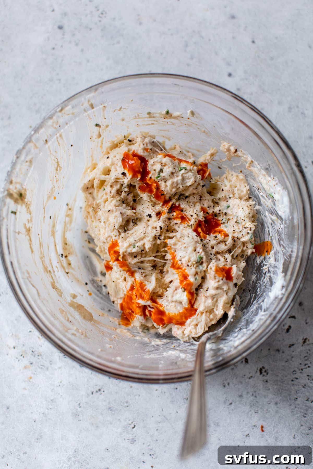 The finished cold crab dip mixture is shown in a clear glass bowl, with a dash of hot sauce freshly added, ready to be stirred in for a customizable level of spicy kick. A spoon rests within the bowl.