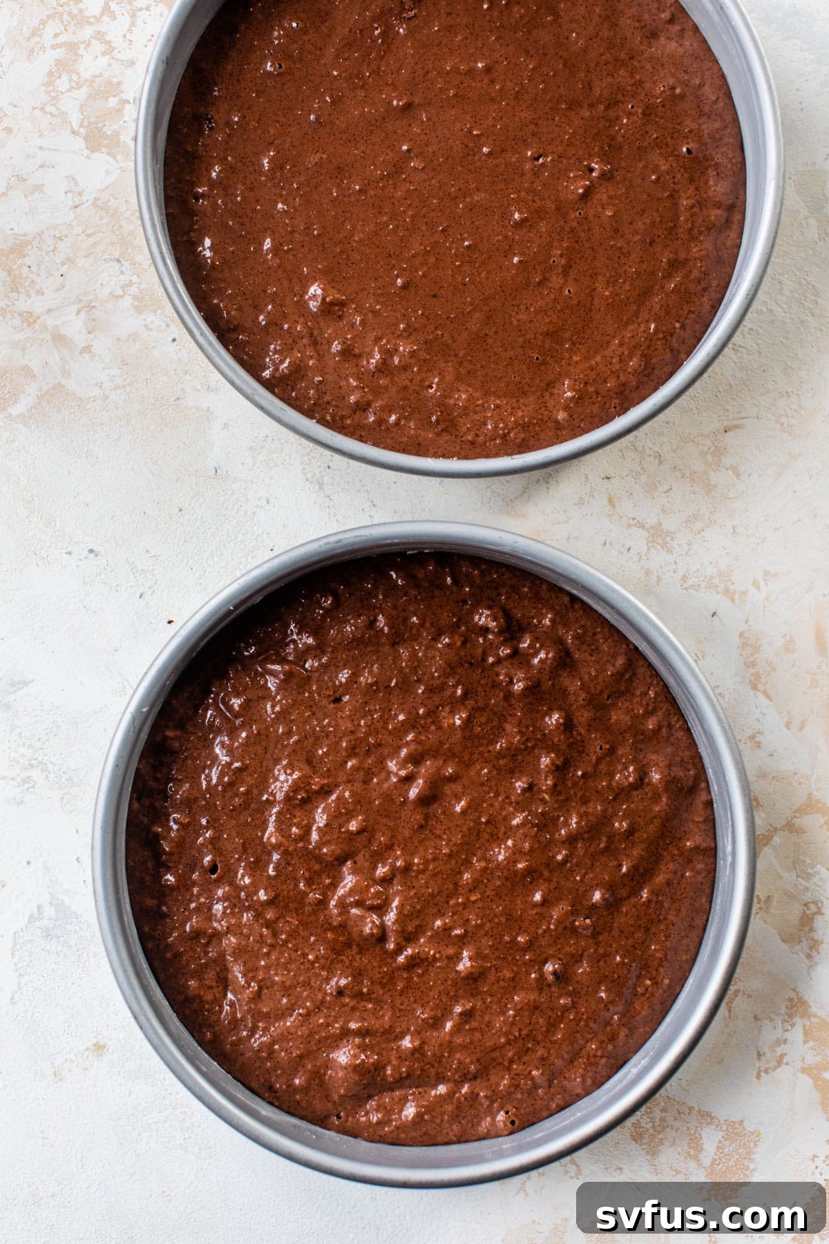 Rich chocolate cake batter evenly divided into two greased and parchment-lined 8-inch round cake pans, ready for baking.