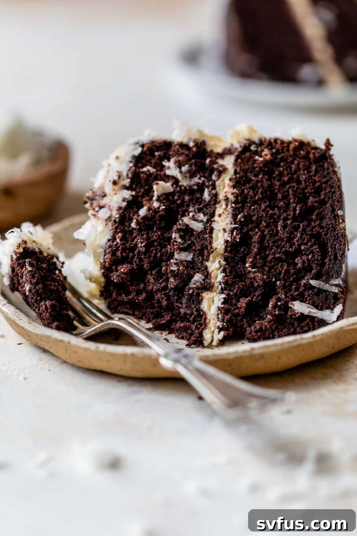 A close-up of a single slice of Chocolate Coconut Cake on a white plate, revealing its inviting layers of dark chocolate cake and smooth white coconut frosting, garnished with shredded coconut, with a fork ready to enjoy.