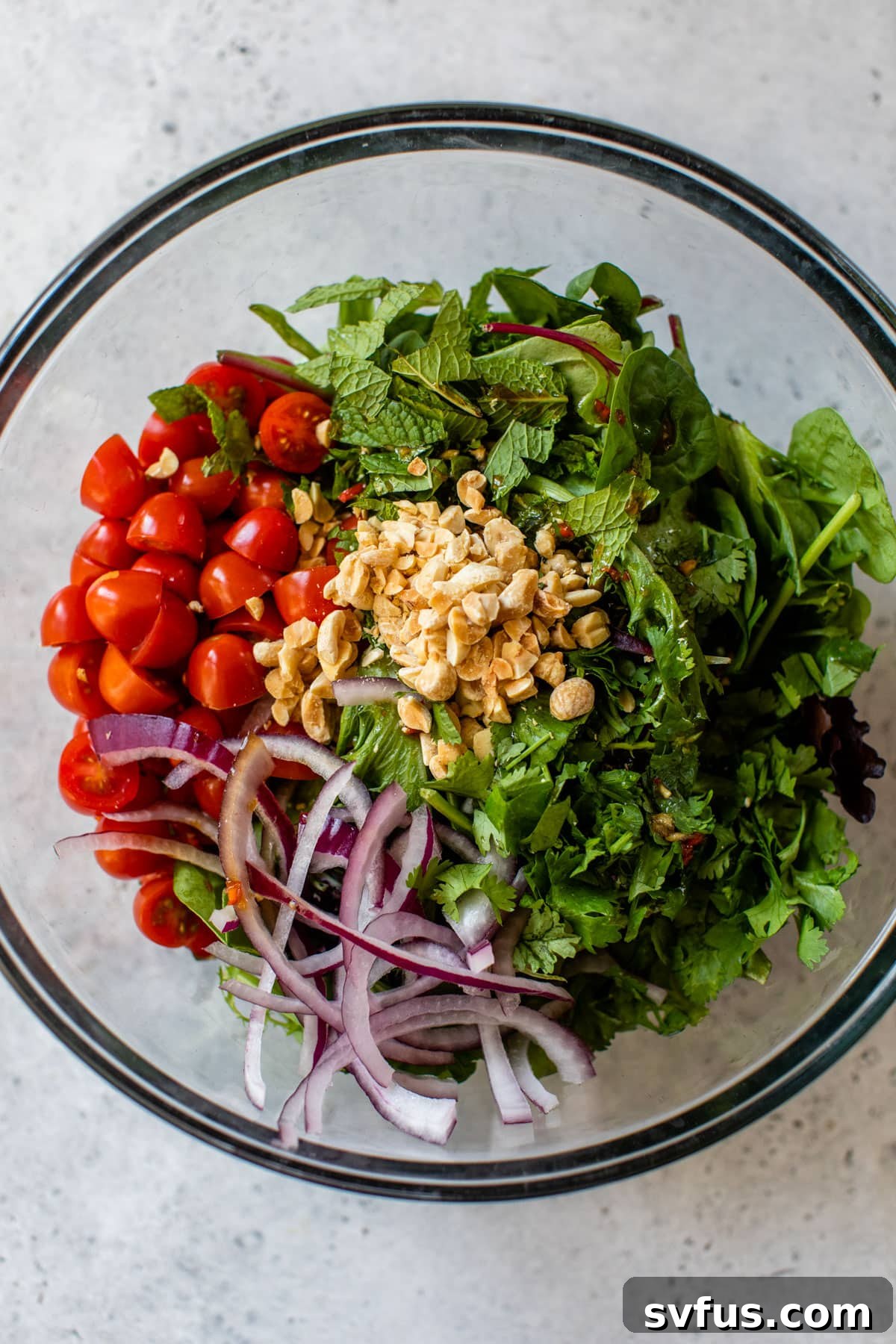 A vibrant mix of fresh vegetables, including mixed greens, grape tomatoes, red onion, cilantro, mint, and chopped peanuts, tossed in a large glass bowl.