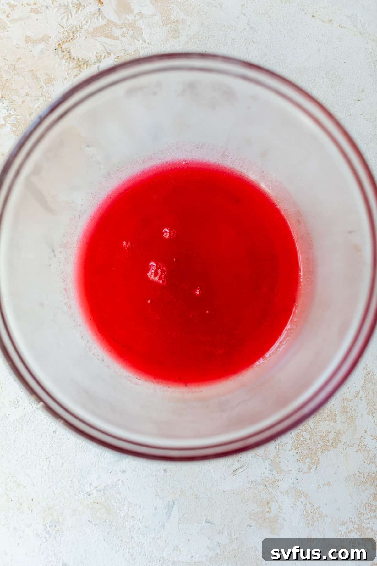 Overhead view of vibrant red cranberry syrup collected in a clear glass bowl after straining.