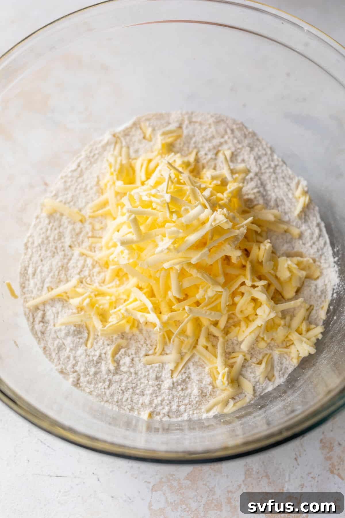 Dry ingredients in a glass bowl, topped with grated cold butter for apple cranberry scones.