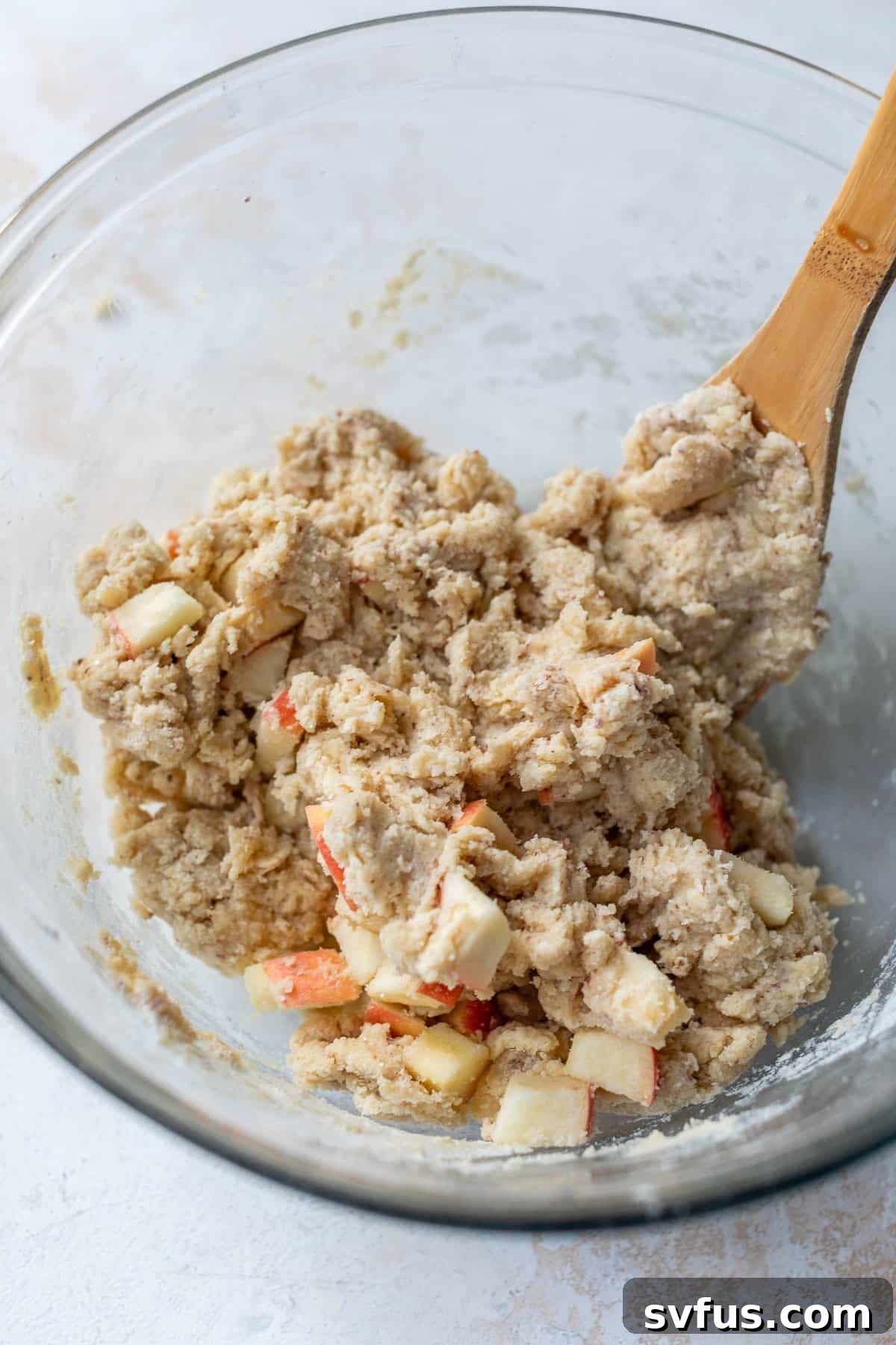 Using a wooden spoon to gently fold chopped apples into vegan apple cranberry scone dough in a glass bowl.