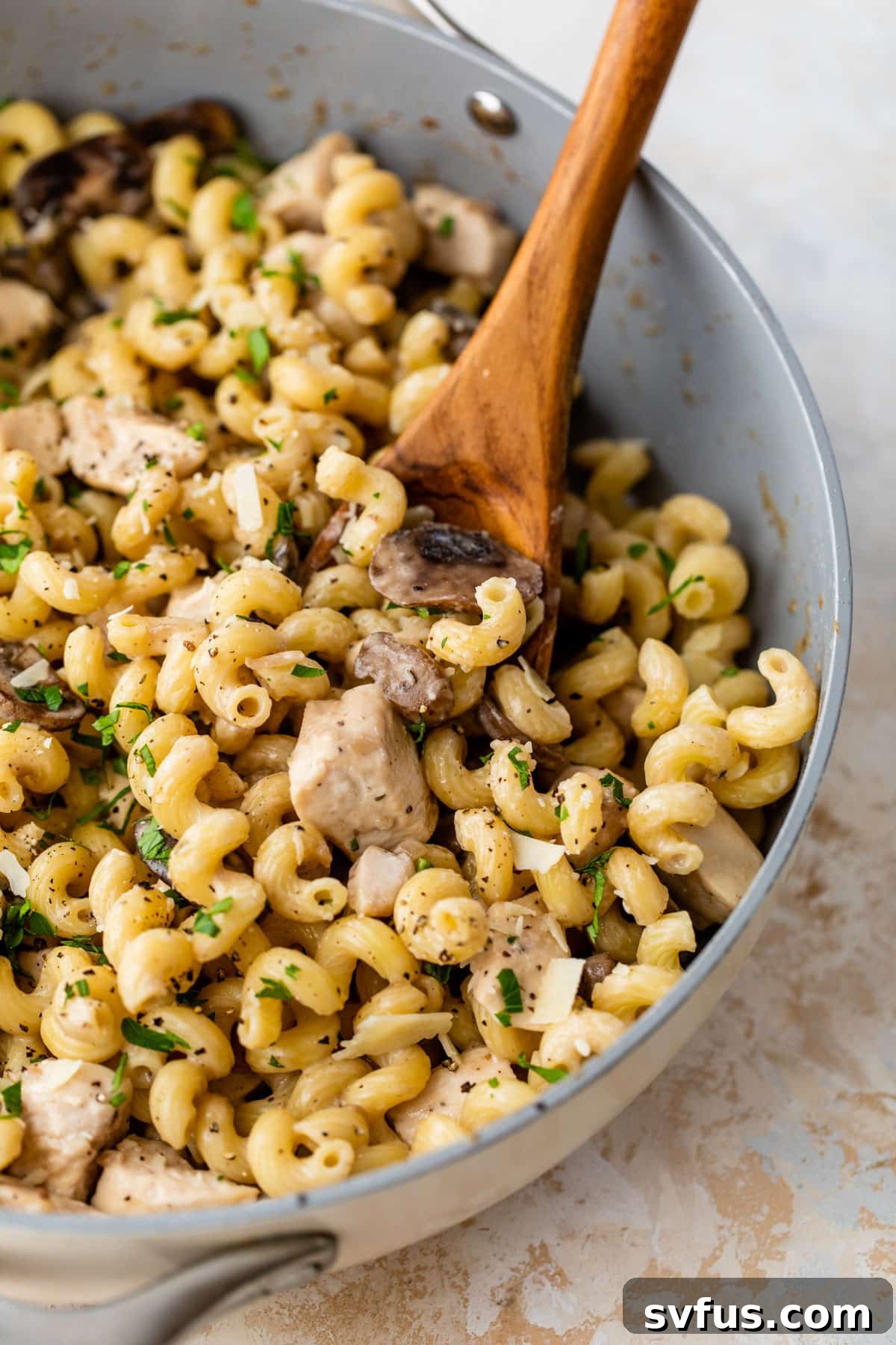 Close-up view of creamy chicken mushroom pasta in a large skillet, ready to be served, with a wooden spoon for stirring.