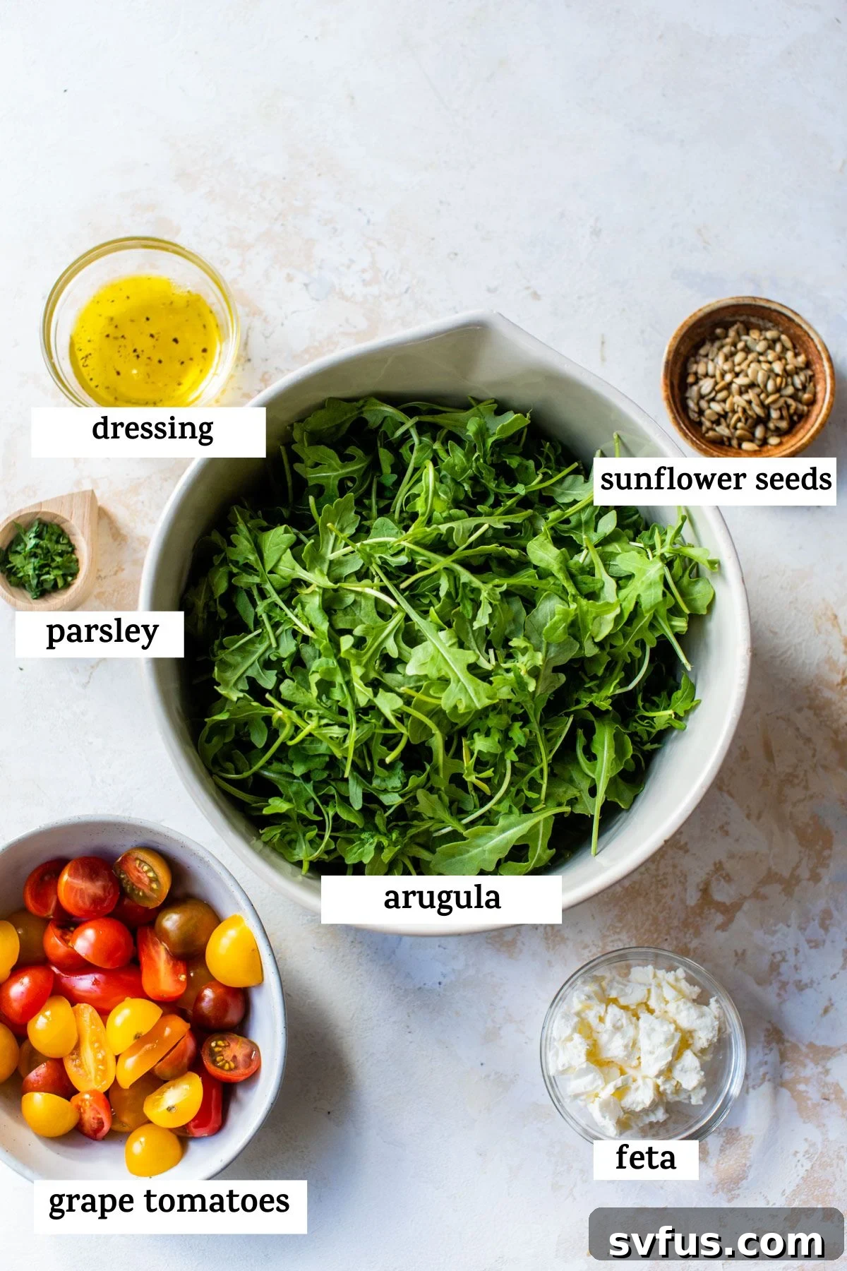 A flat lay of fresh salad ingredients artfully arranged on a rustic wooden table: a bowl of vibrant dressing, a sprinkle of sunflower seeds, fresh green parsley, a generous bed of arugula, bright red grape tomatoes, and a block of creamy feta cheese.