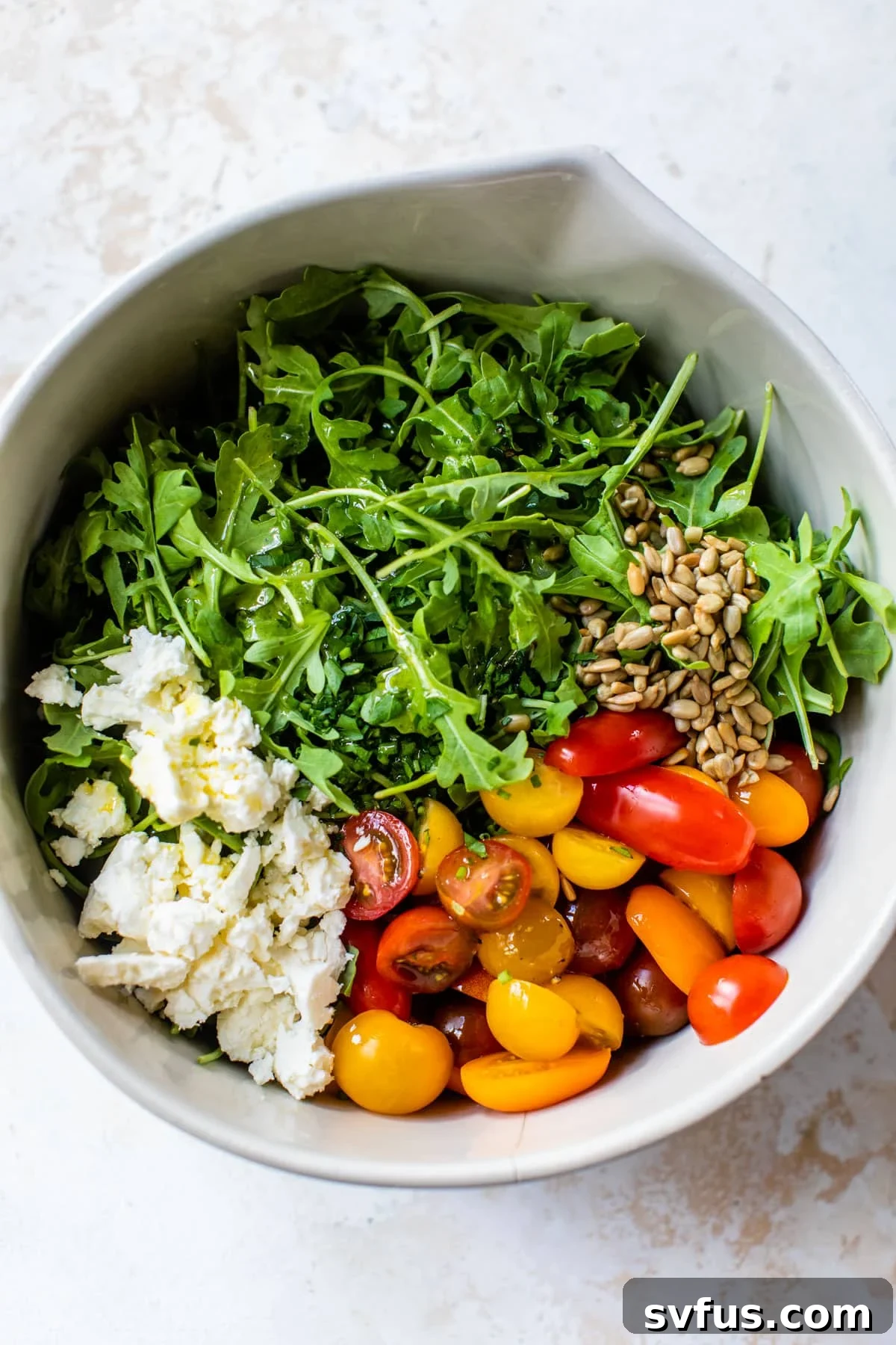 A large mixing bowl filled with fresh arugula salad ingredients before dressing: vibrant green arugula, sliced red grape tomatoes, crumbled white feta cheese, and a sprinkle of sunflower seeds.