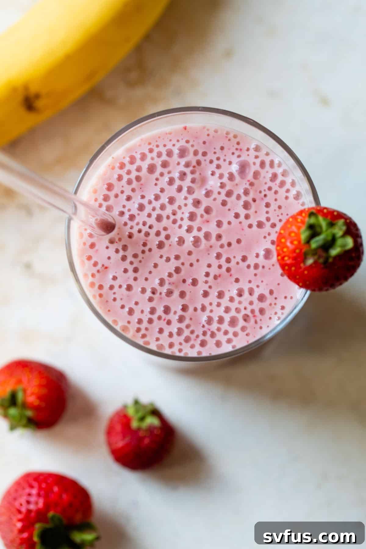 Close-up view of a strawberry banana smoothie in a clear glass, with a colorful straw and a strawberry resting on the rim.
